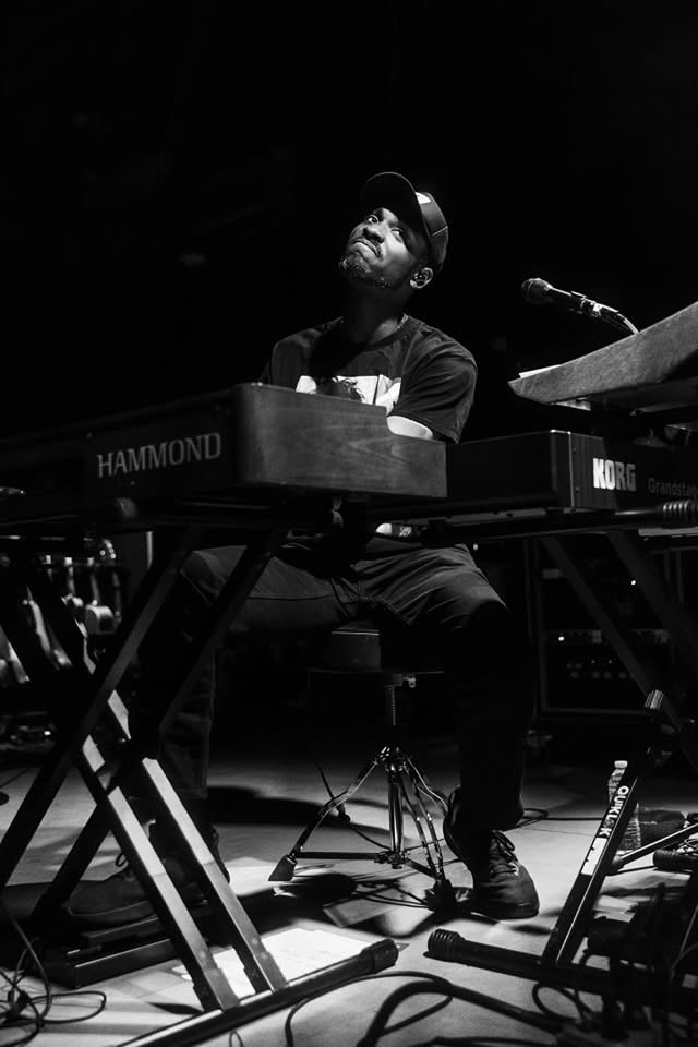 Person playing keyboard on stage, wearing a cap and glasses, with a microphone nearby, in black and white.
