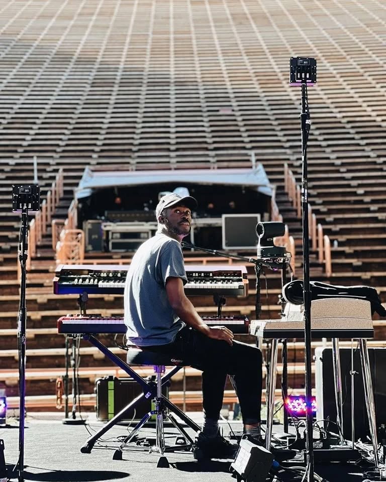 Person sitting at a keyboard on stage with musical equipment, empty stadium seats in background, wearing a cap and glasses.