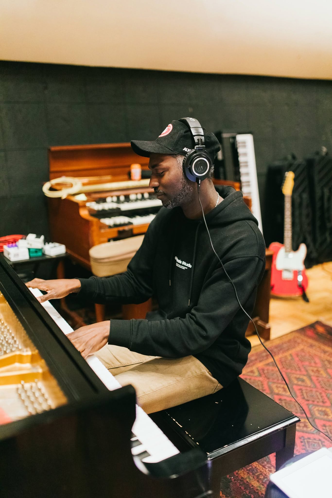 Person wearing headphones and a cap playing piano in a music studio with instruments and equipment in background.