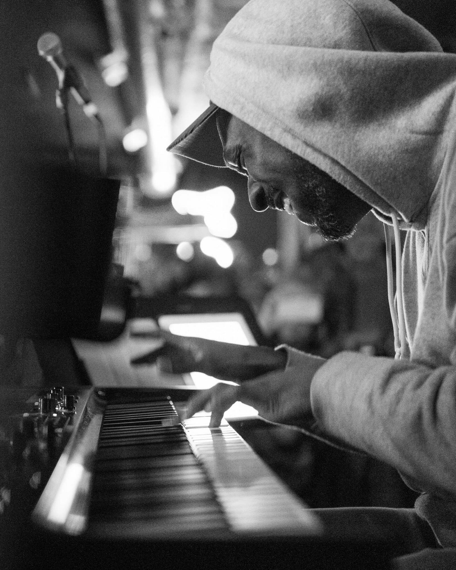 Man wearing hoodie playing piano outdoors at night, focused on keys, with blurred background lights.