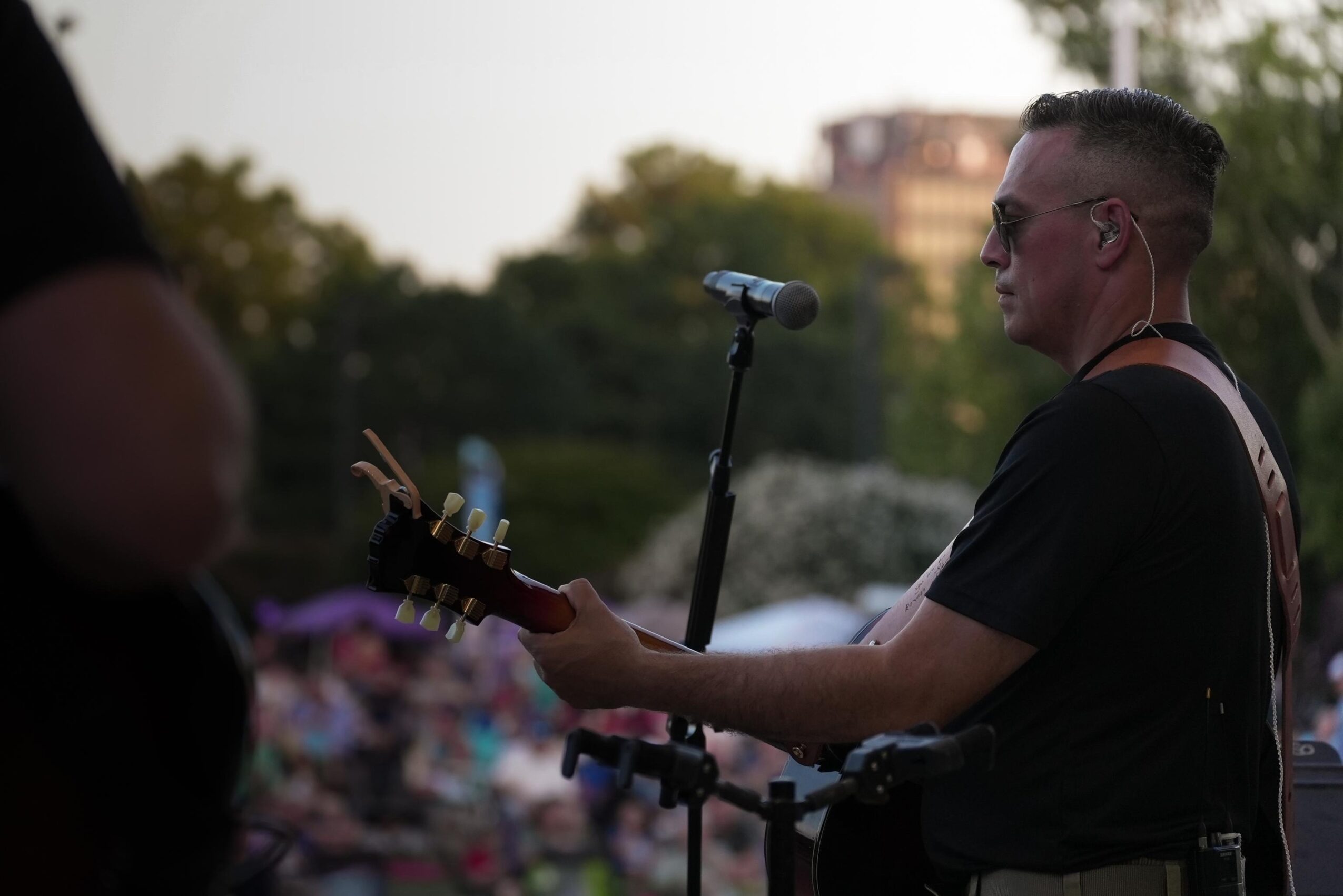 Man playing guitar and singing into a microphone outdoors during daytime, with a crowd and trees in the background.