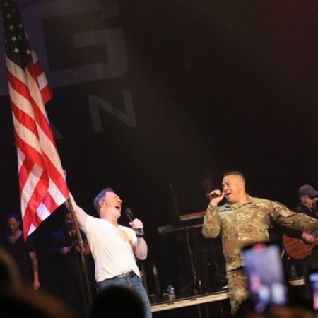 Person in military uniform singing on stage with others, American flag visible, audience taking photos.