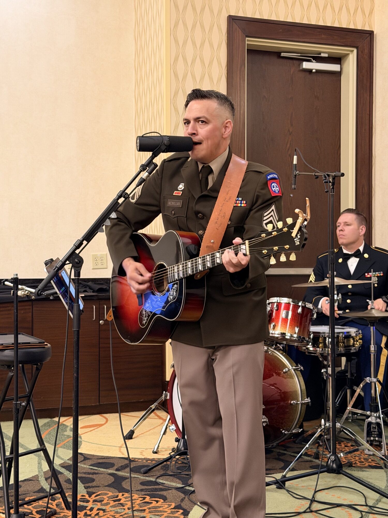 Man in military uniform playing guitar and singing into microphone, drummer in background, indoor setting with beige walls and wooden door.