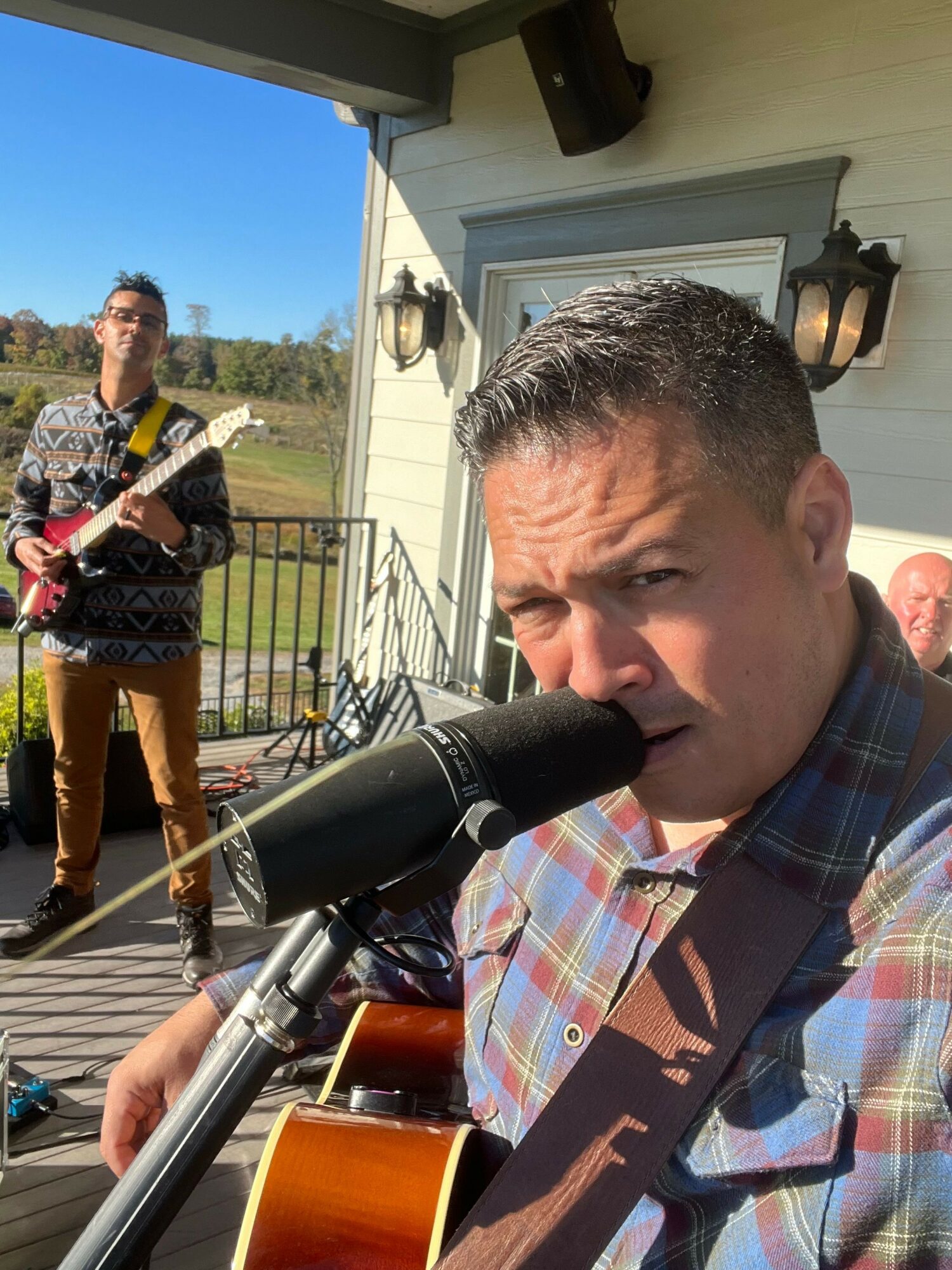 Man playing guitar and singing into a microphone outdoors on a sunny day.
