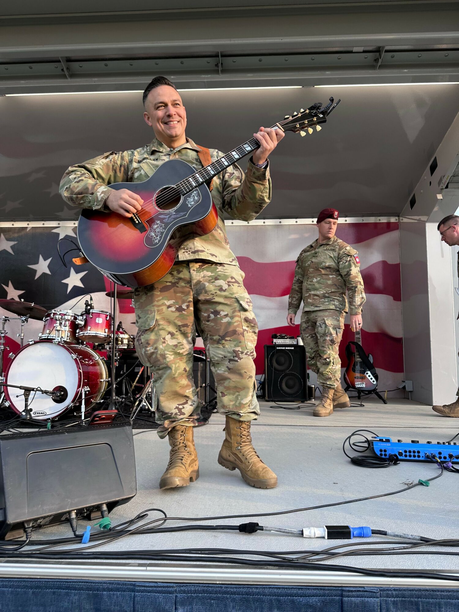 Person in military uniform playing guitar on stage with drum set and equipment, two other people in background, stage setup.