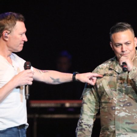 Two men on stage, one pointing at the other, both holding microphones, with a dark background.