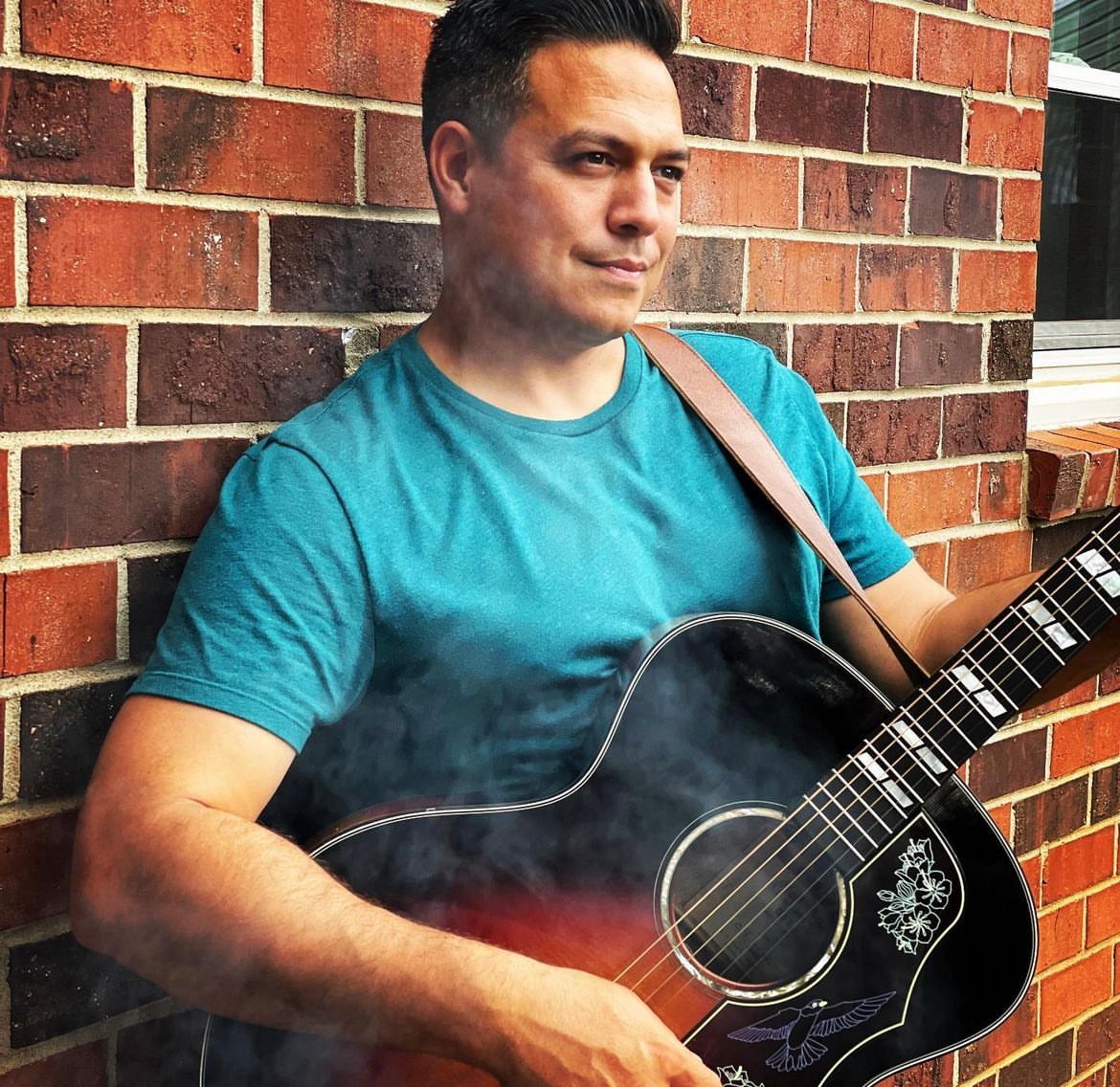 Man with short dark hair wearing a teal T-shirt, holding a black acoustic guitar, standing against a brick wall.