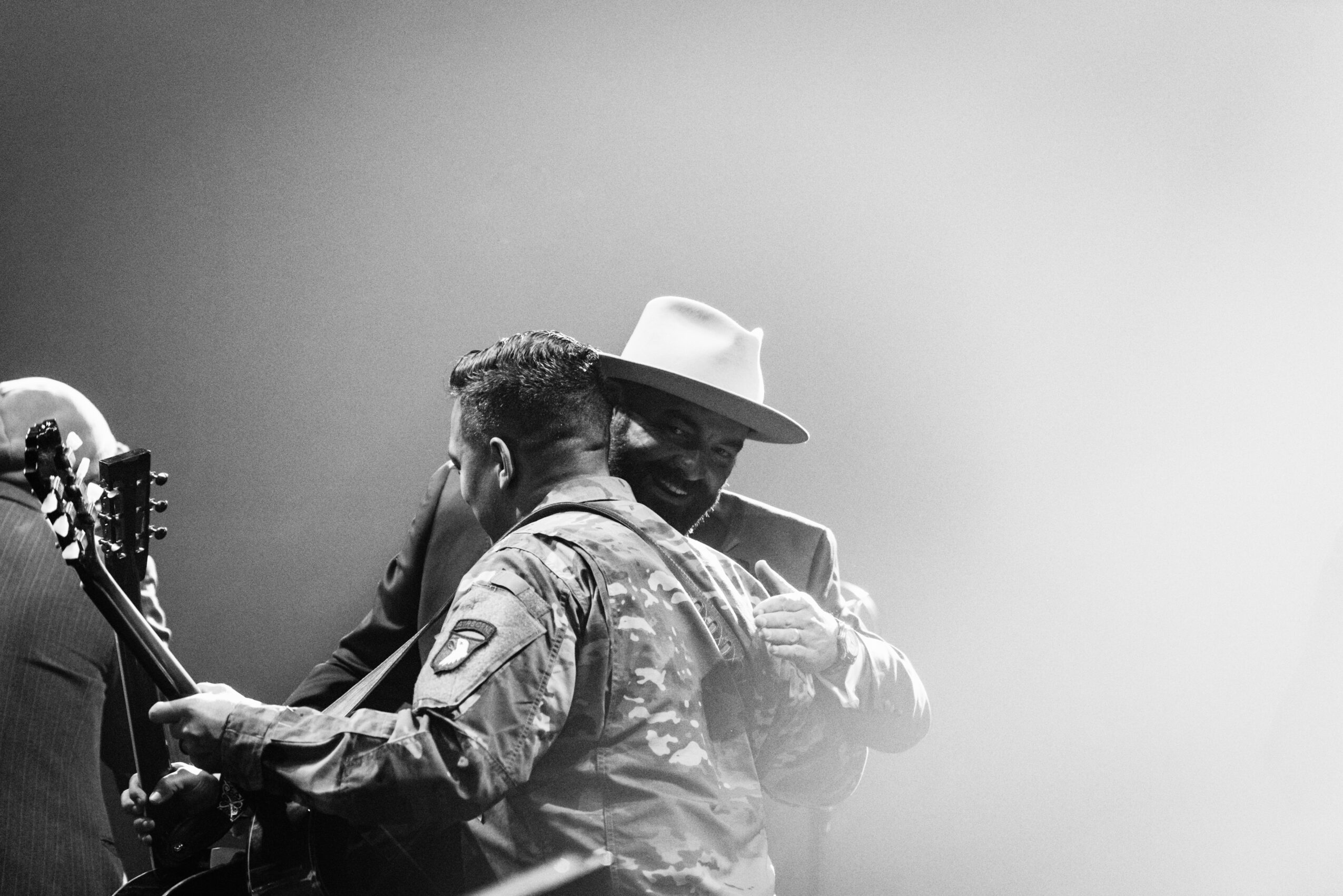 Two men, one in military uniform and the other in a hat, embrace on stage with a guitar nearby, black and white image.