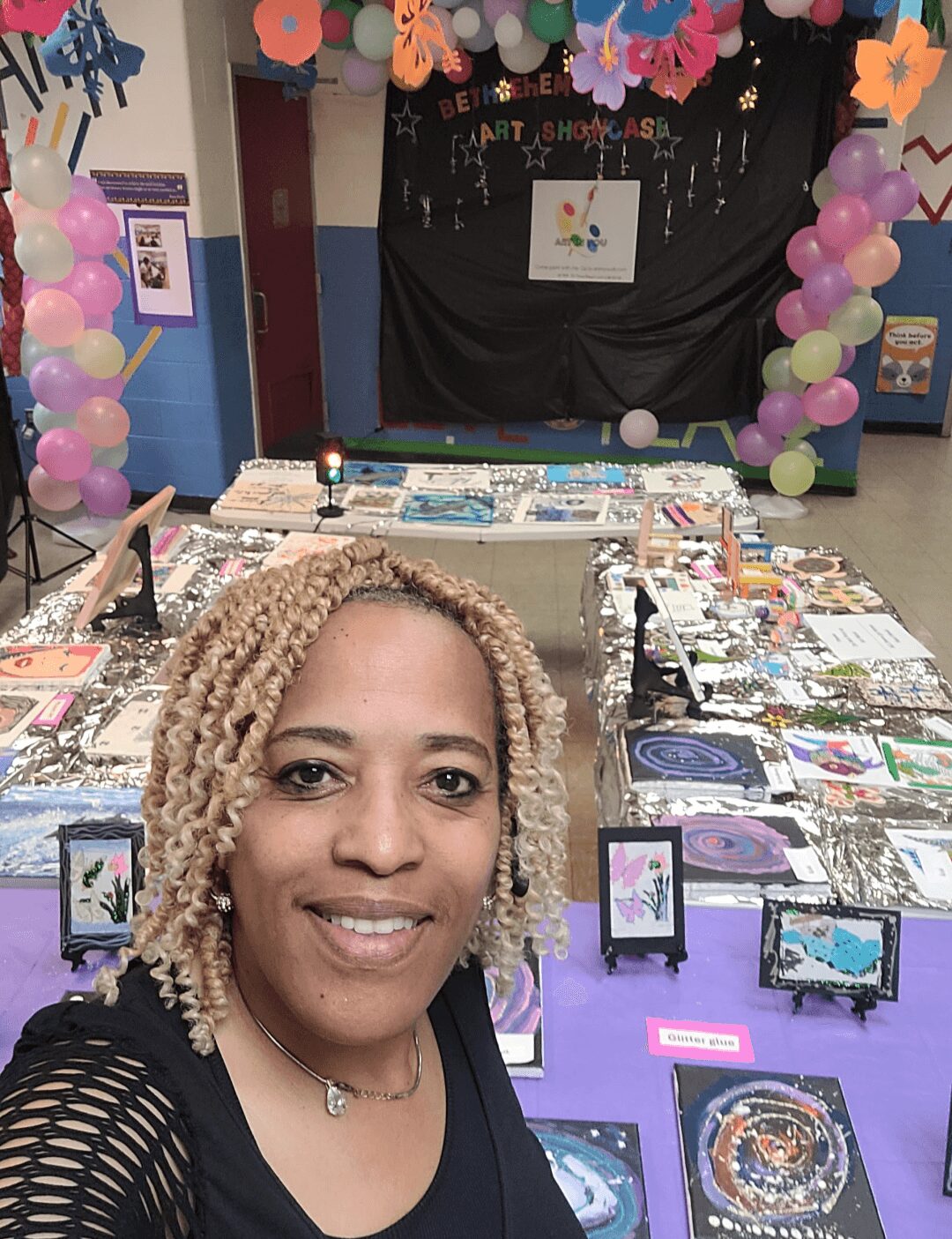 Woman with blonde curly hair smiling at a decorated event table with books and artwork, colorful balloons and decorations in background.