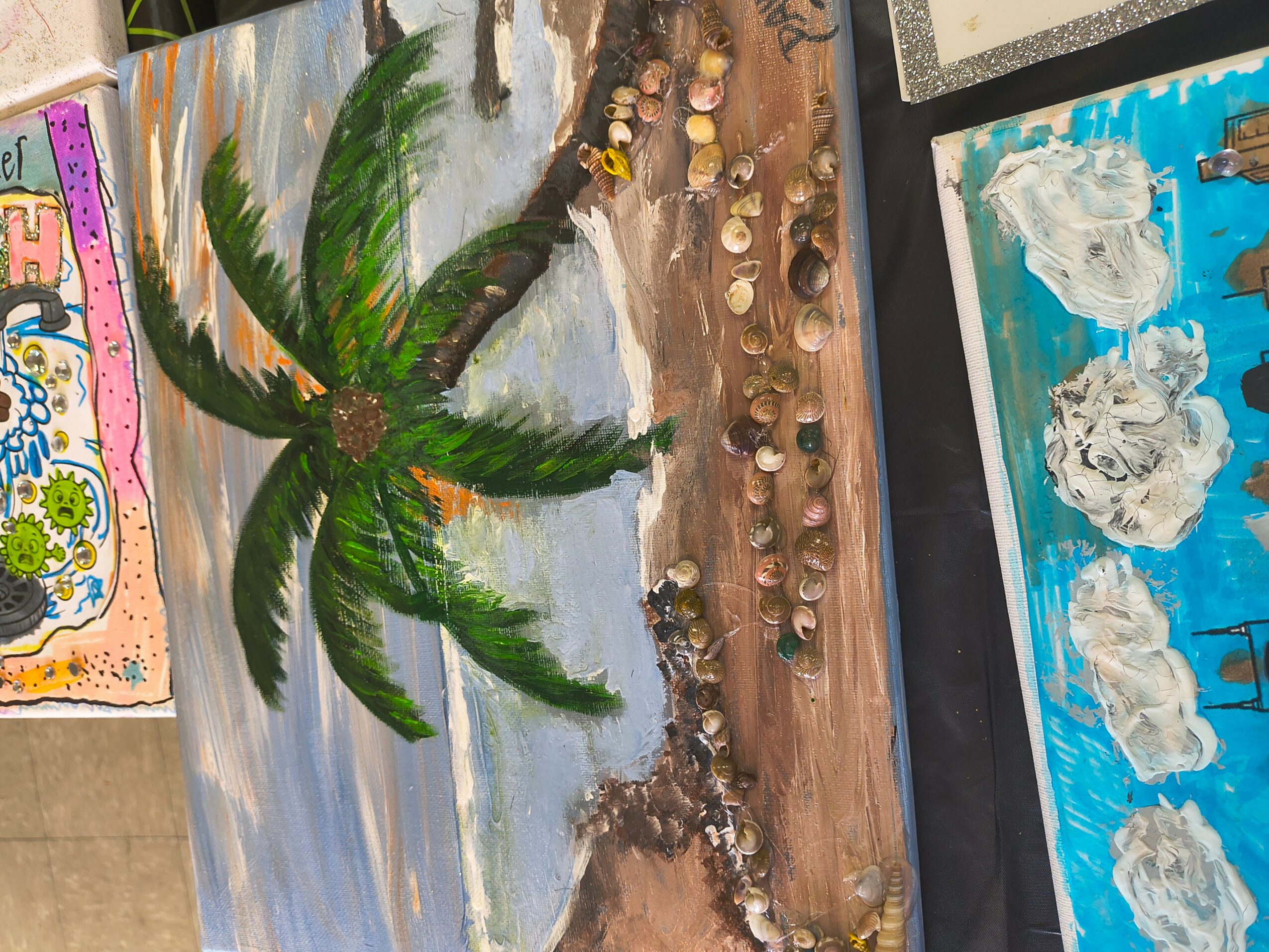 Painting of a palm tree on a beach with sand and water, surrounded by other artwork.