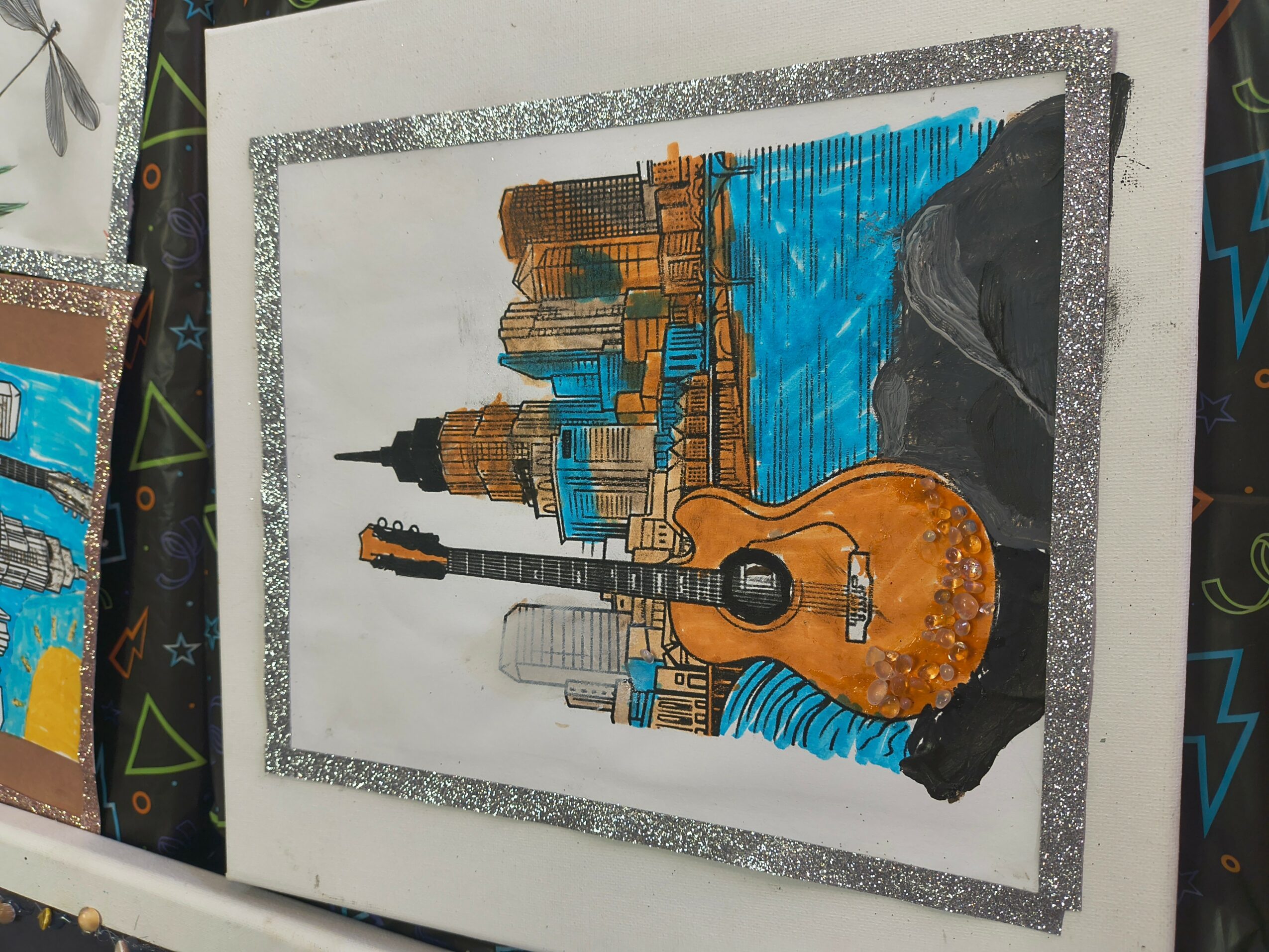 Painting of a city skyline with a guitar in the foreground, water, and rocks, framed with glittery border.