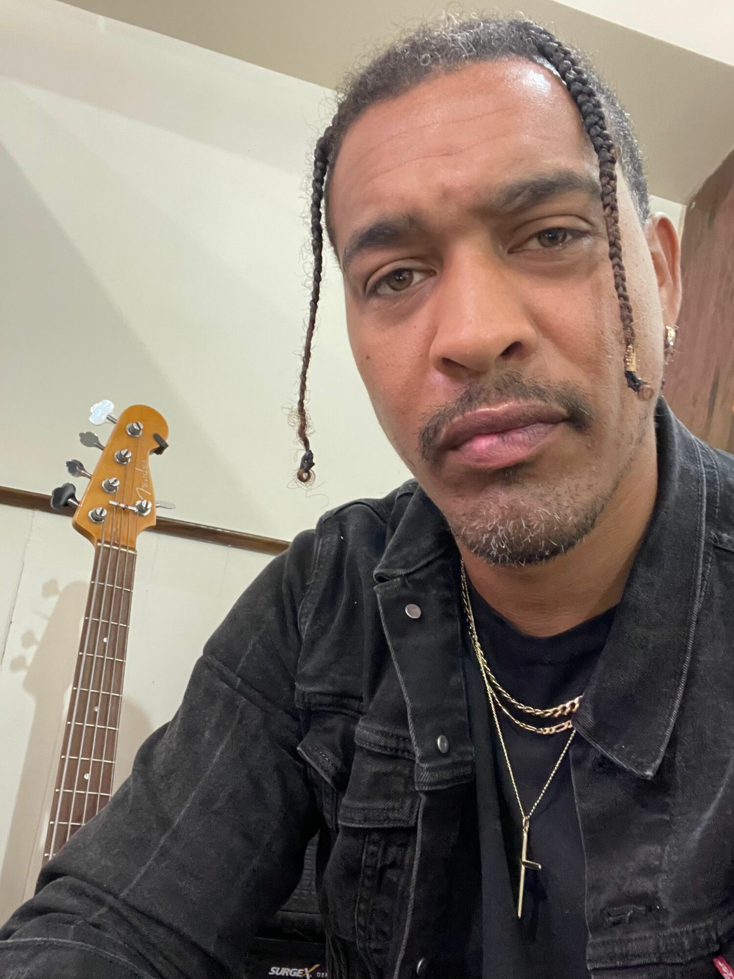 Person with braided hair and facial hair, wearing a black jacket, sitting near a guitar, looking at the camera.