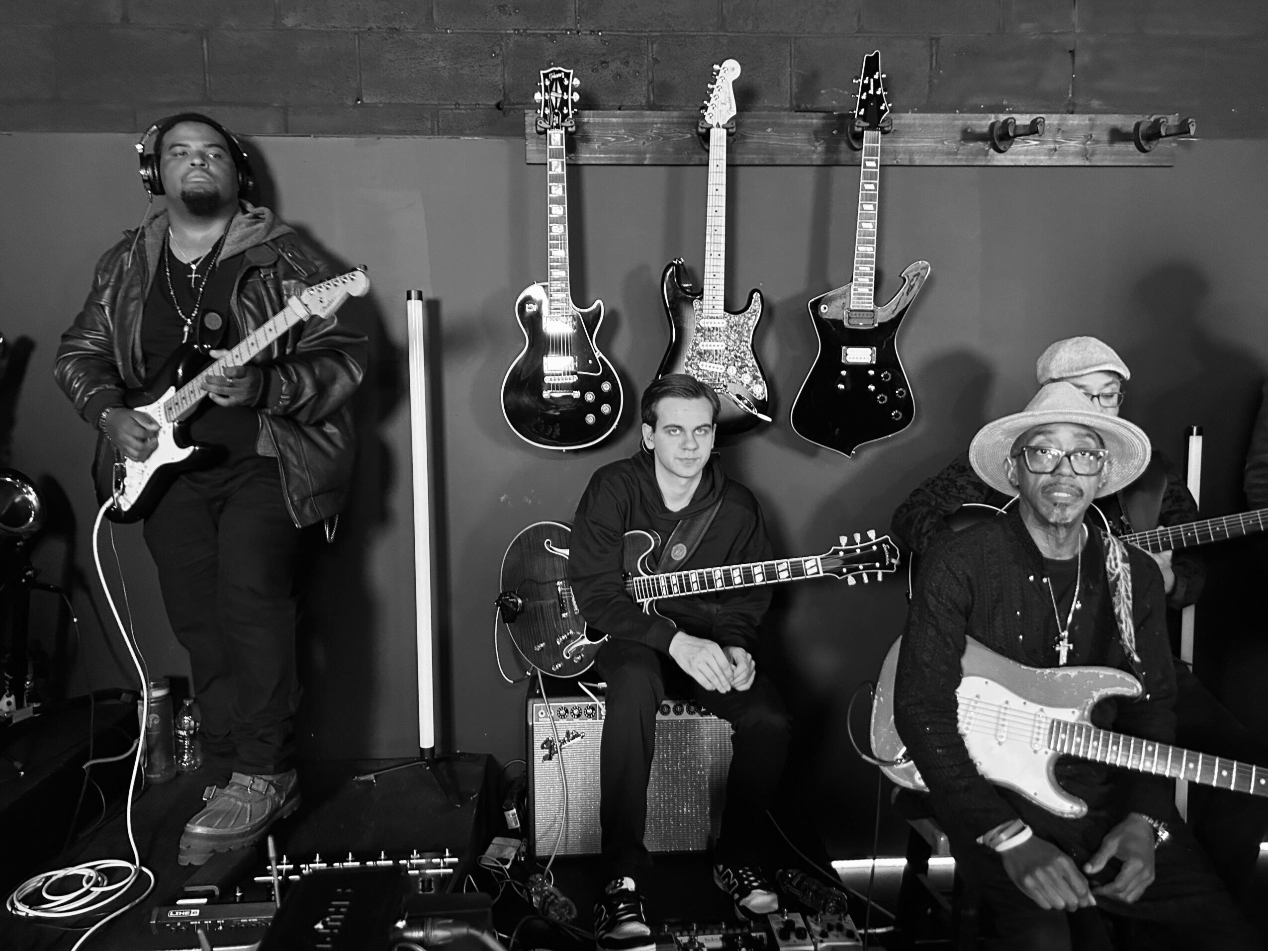 Four musicians with guitars, one sitting, one standing, in a room with hanging guitars on the wall.