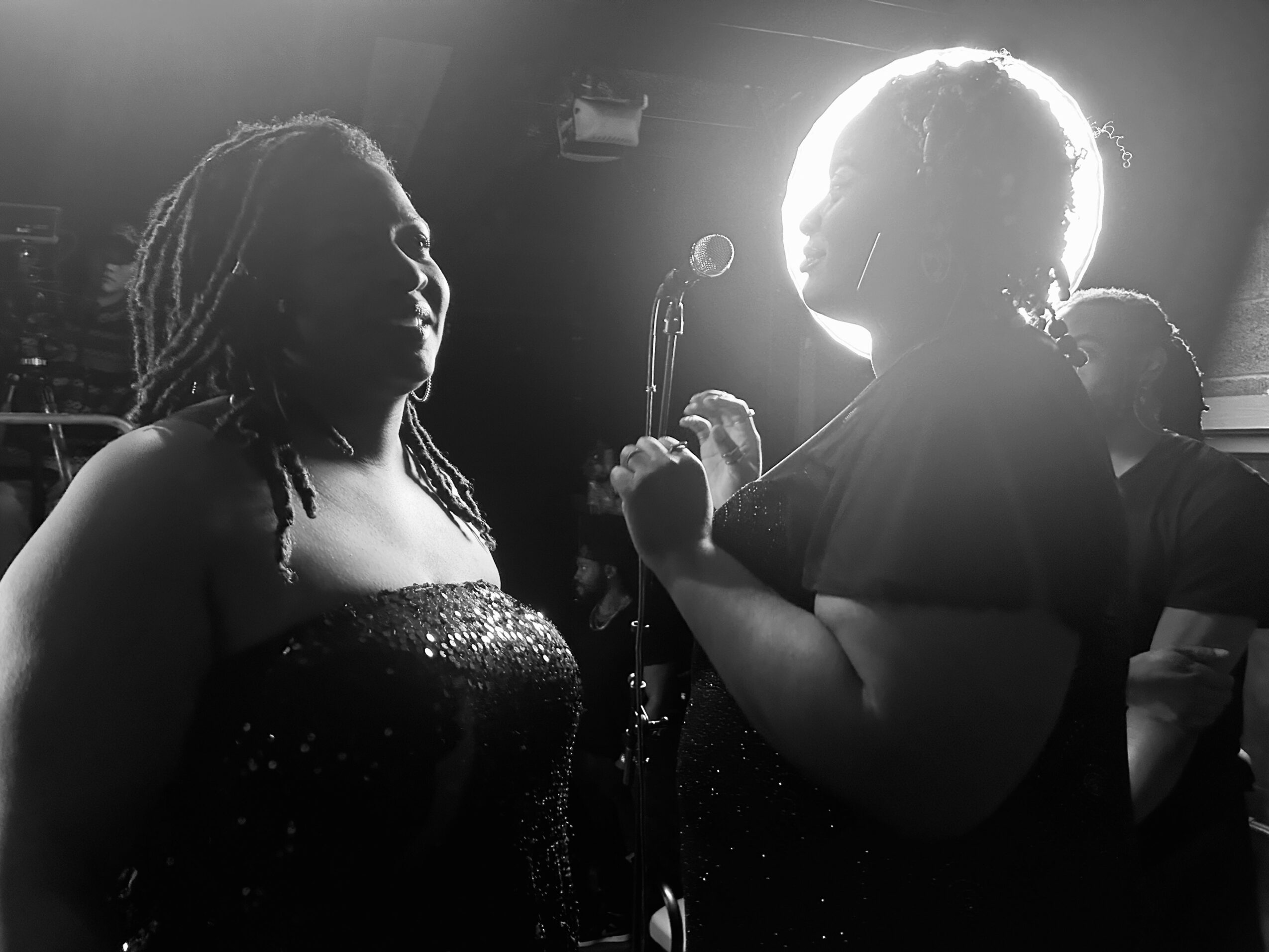 Two women face each other on stage, one holding a microphone, with a bright light behind the other, in black and white.