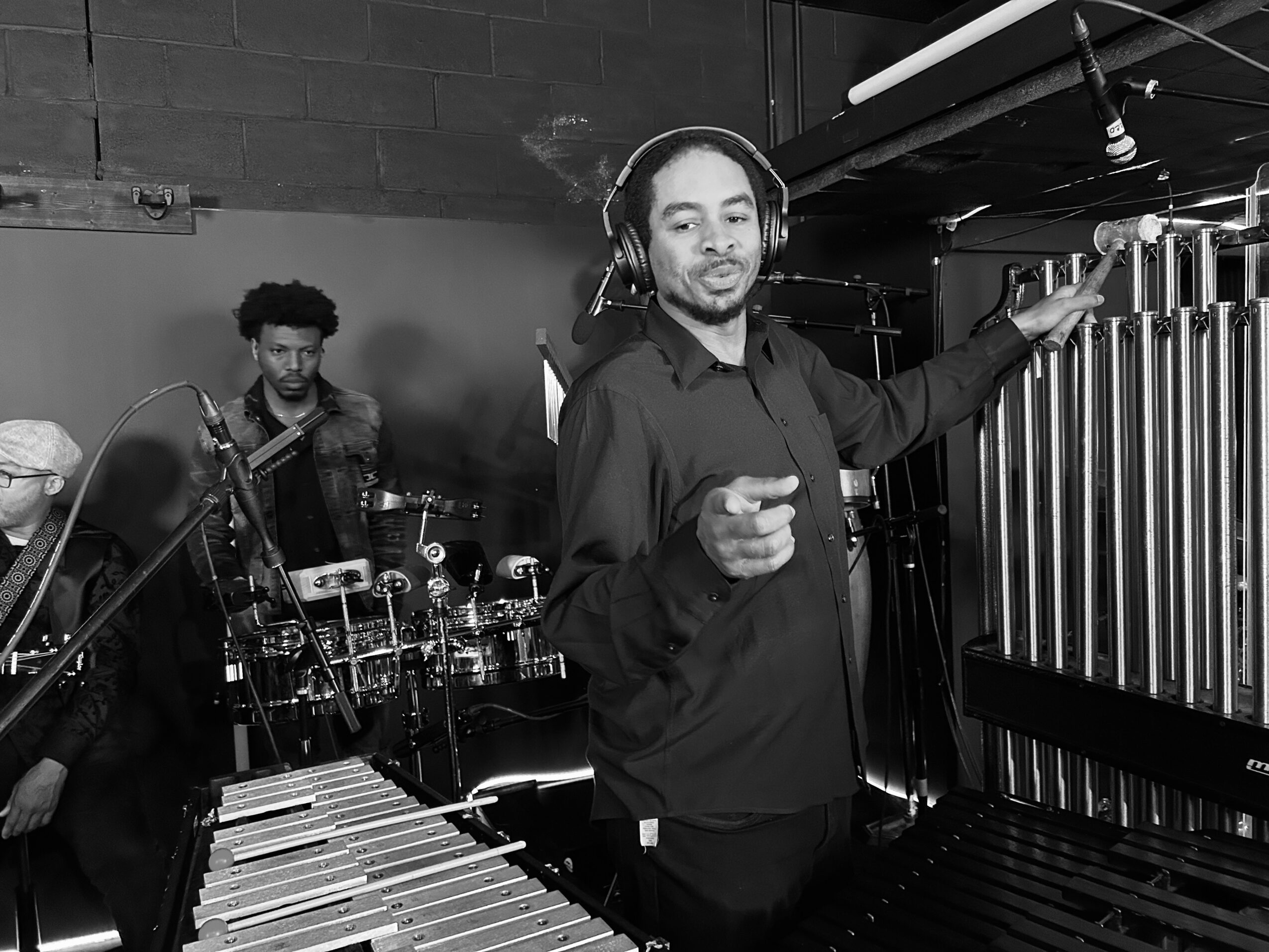 A man with headphones stands in front of a vibraphone, pointing at it, with a band playing in the background.