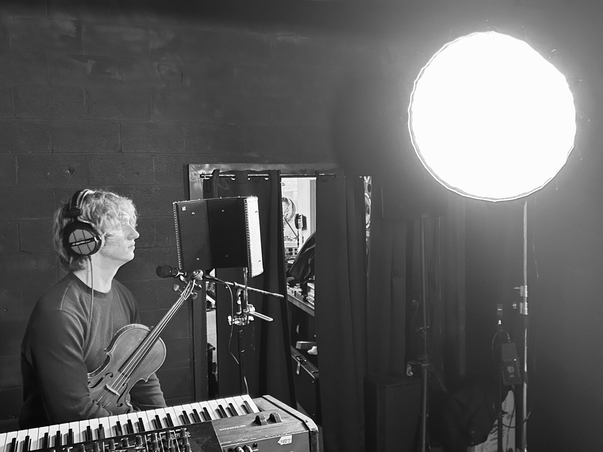 Person with headphones holding a violin in a recording studio, with a keyboard and a large light source nearby.