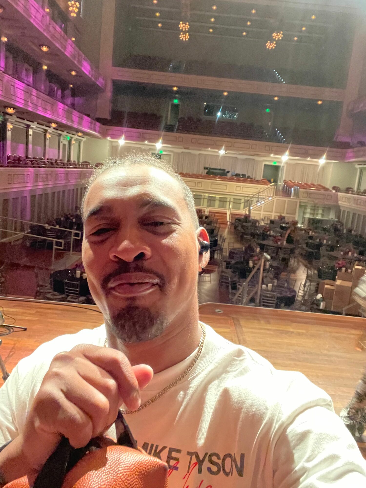 Person with short hair making a funny face, holding a camera or phone, in a brightly lit indoor space.