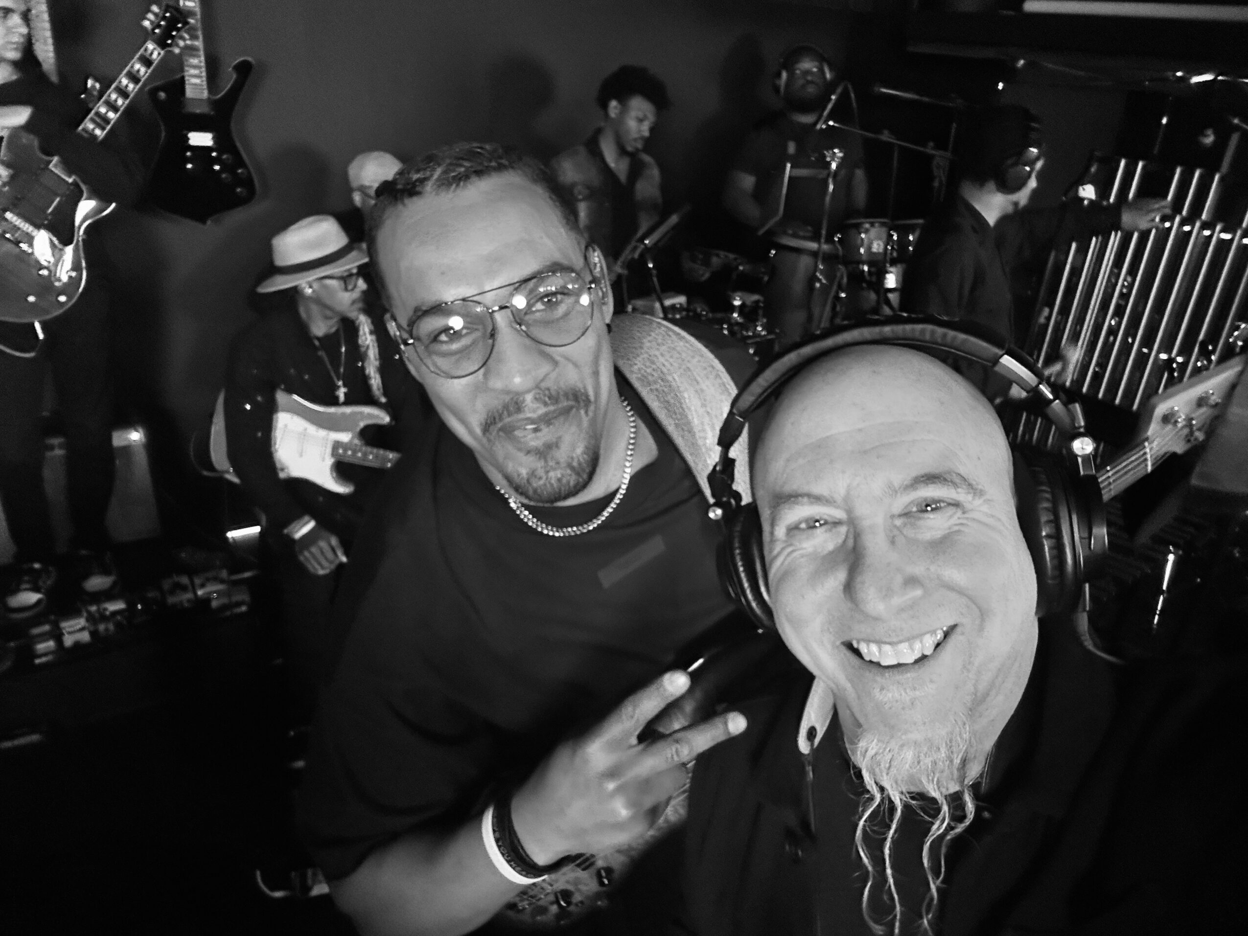 Two men smiling, one making a peace sign, in a music studio with band members and instruments in background.