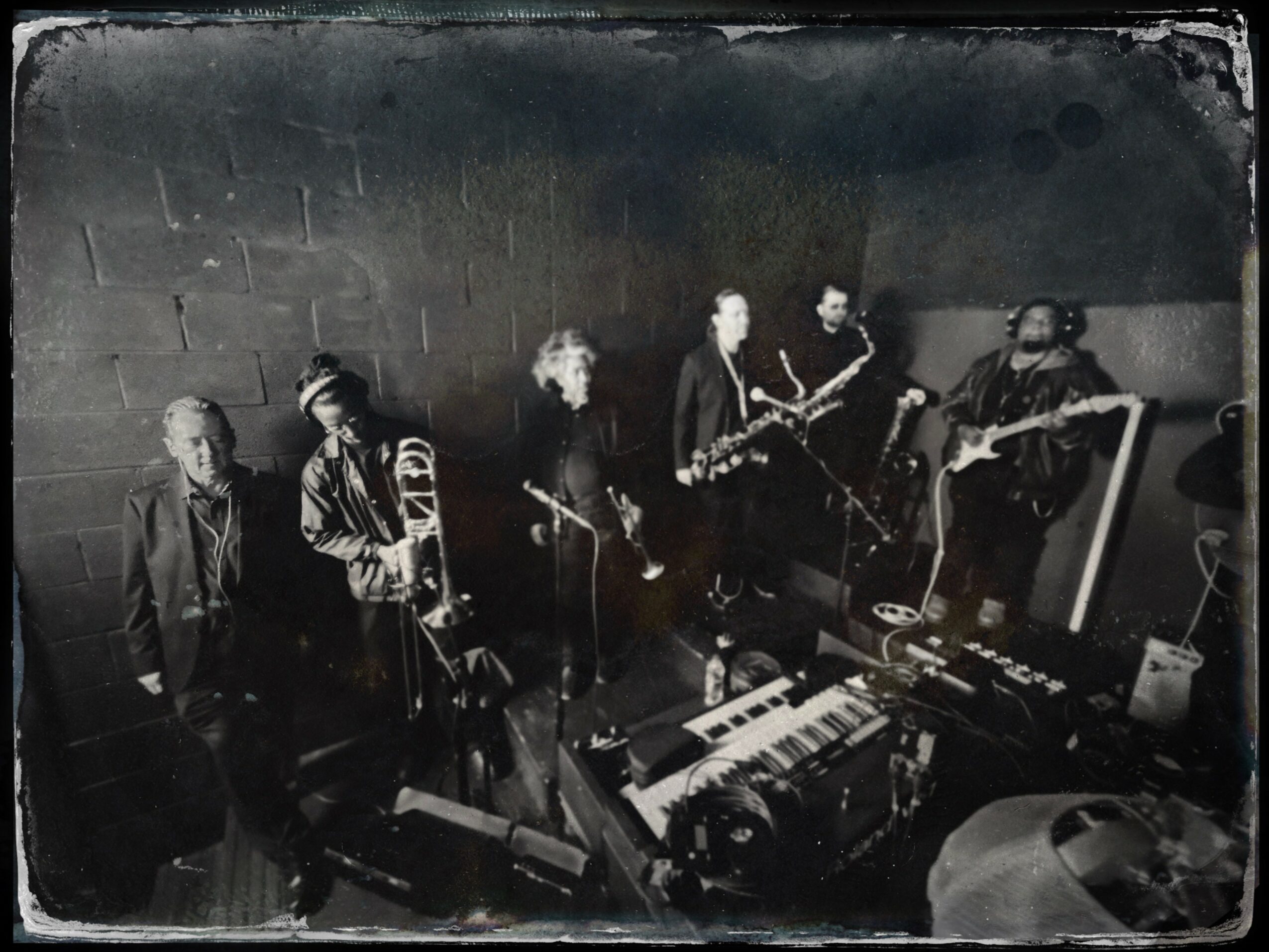 Group of musicians with instruments in a dimly lit room, including keyboard and saxophone, with a brick wall background.