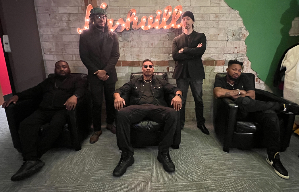 Five people pose in a room with a brick wall and neon sign, some sitting and some standing, dressed in dark clothing.