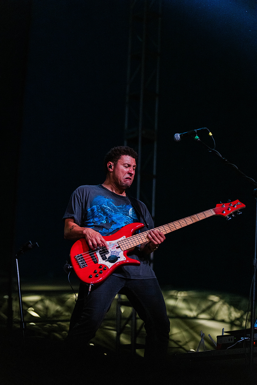 Musician playing a red electric guitar on stage with dark background and microphone nearby.