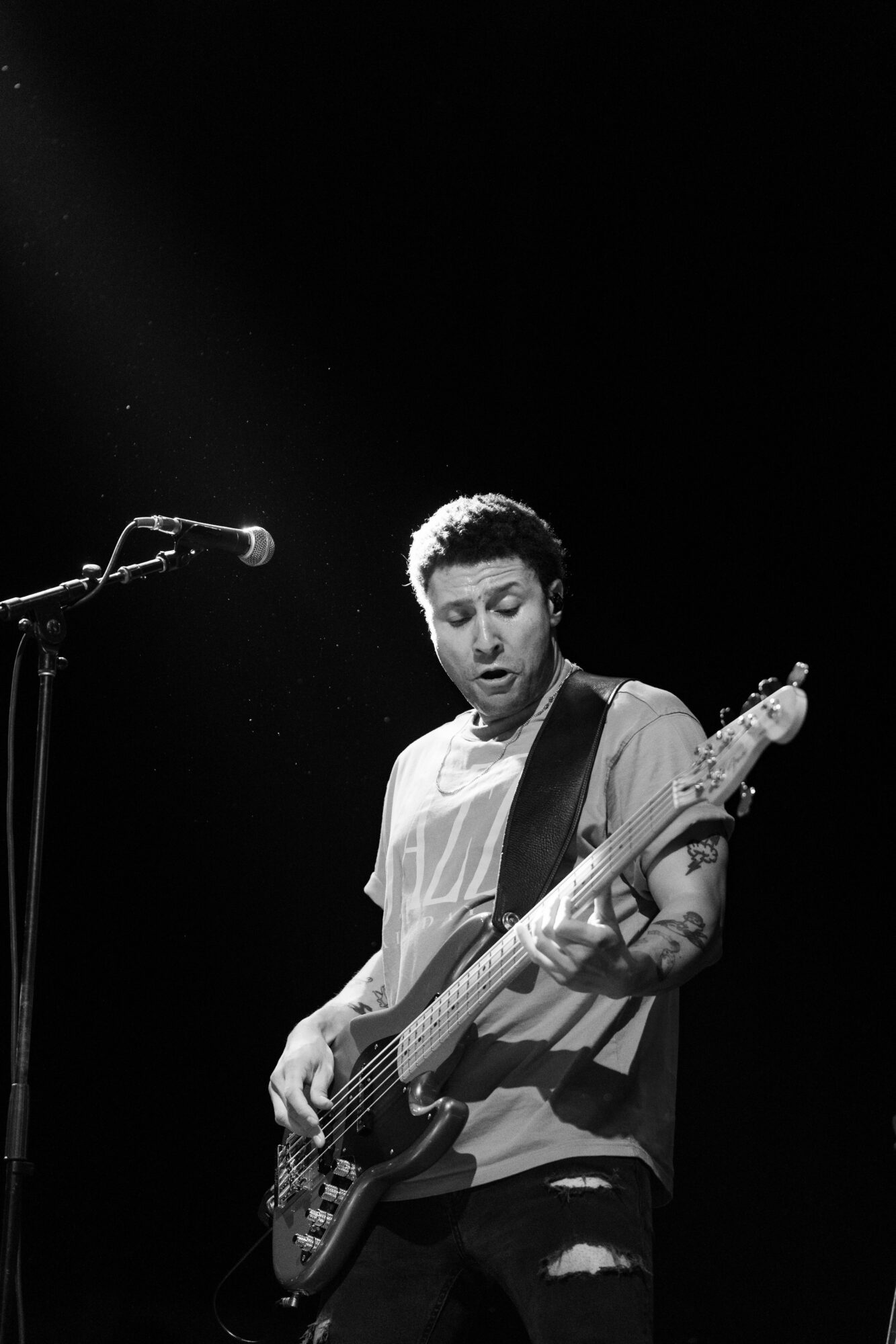 Person playing bass guitar on stage with microphone, black background, focused expression, short curly hair, wearing a light-colored shirt.