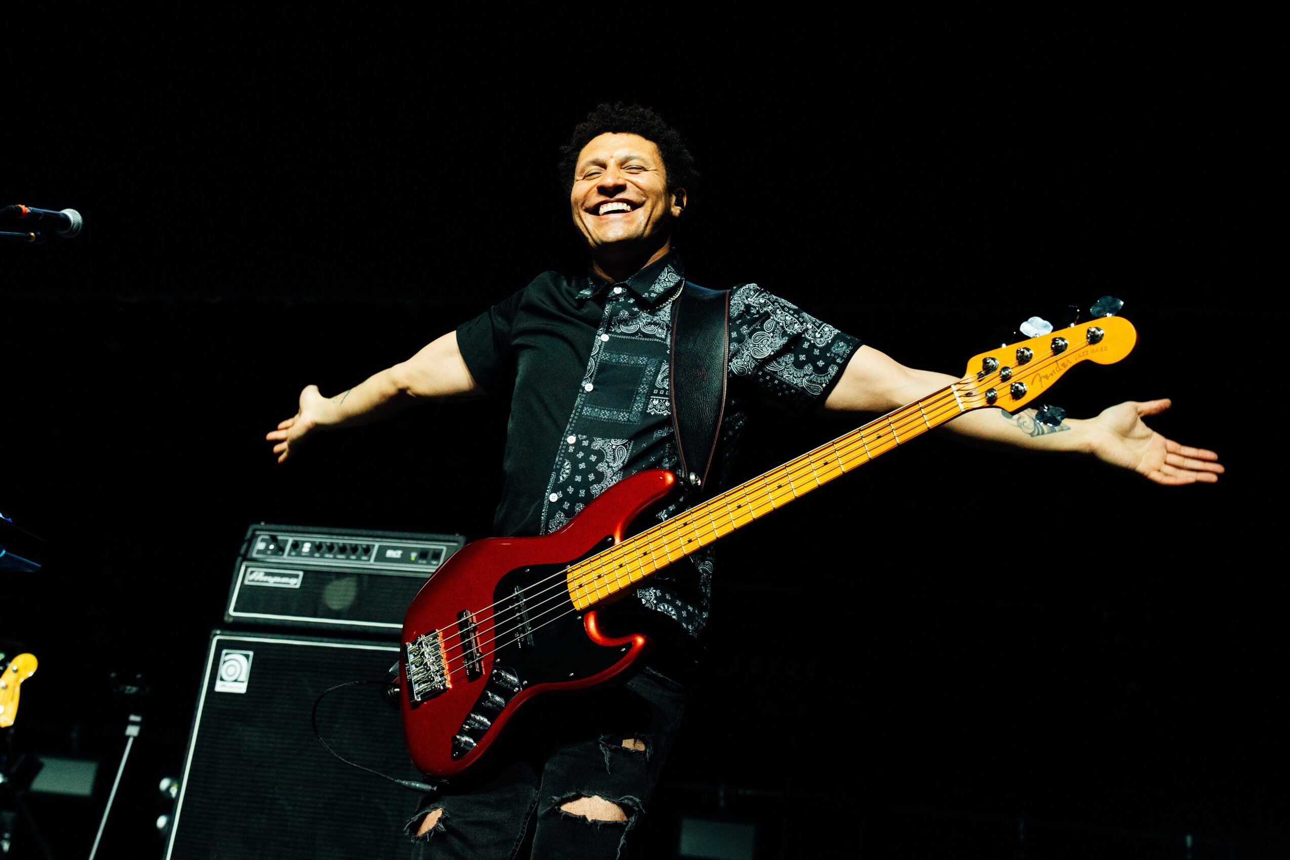 Person smiling with arms outstretched, holding a red bass guitar on stage, with musical equipment behind them.