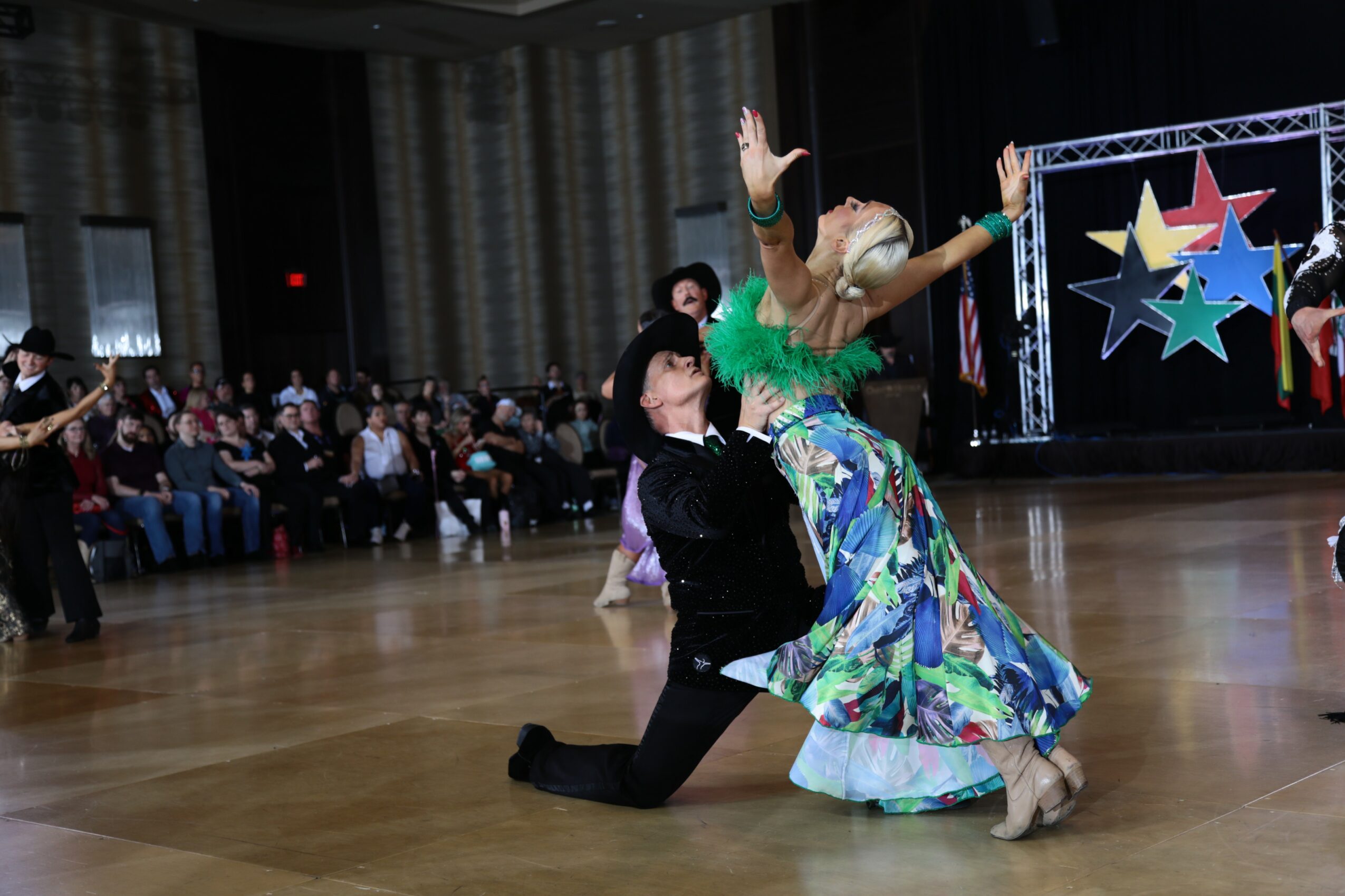 Two dancers perform on a wooden floor with an audience watching, colorful stage backdrop with stars, one dancer in a floral dress, the other in black.