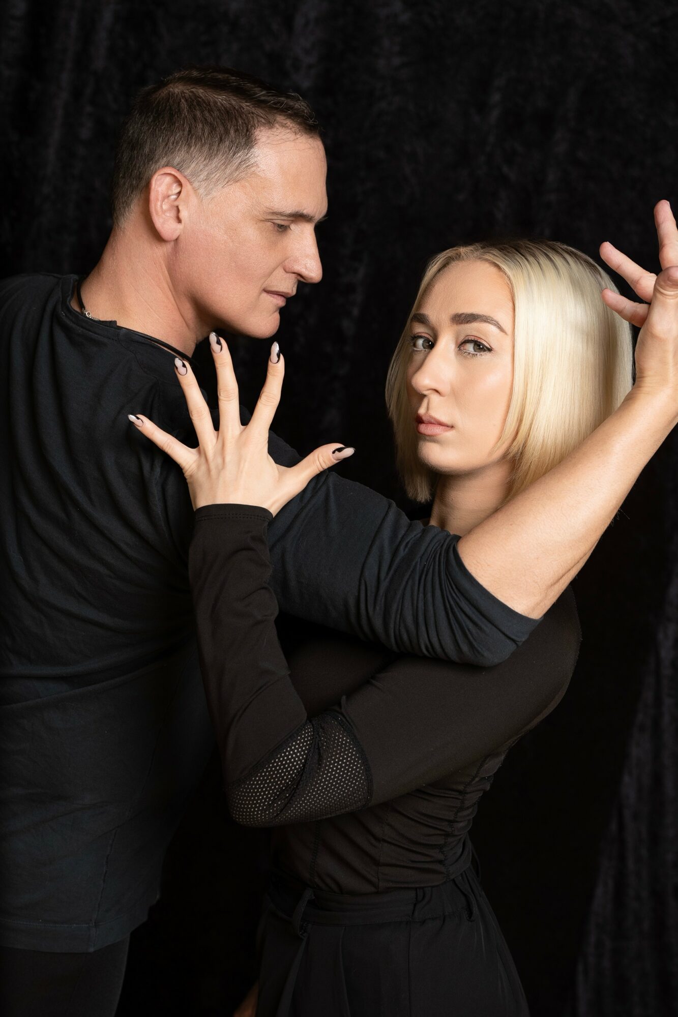 A man and woman pose closely with serious expressions, hands raised and touching each other, against a dark background.