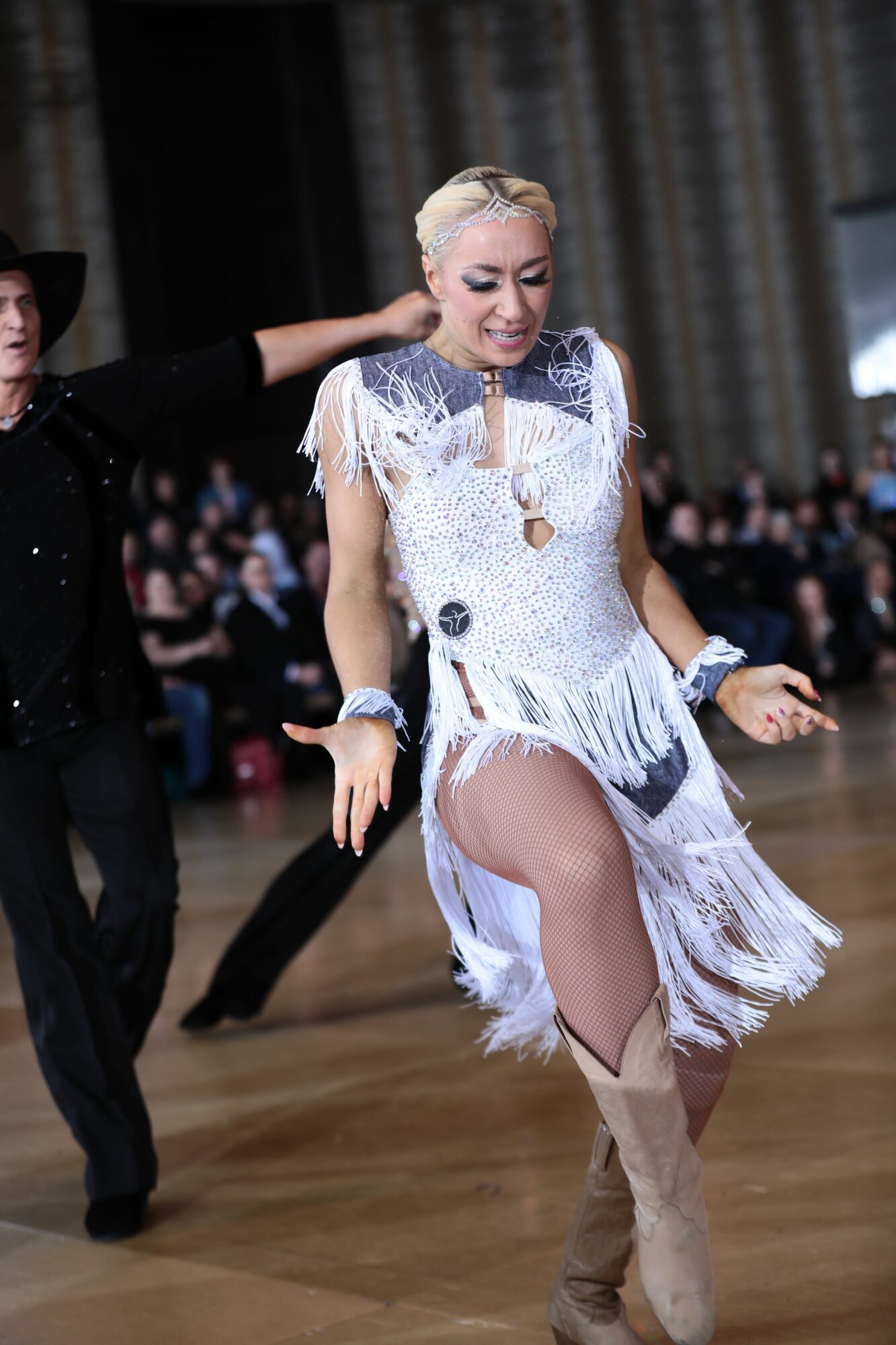 A woman in a white dress with fringe dancing on a wooden floor, with a man in black behind her and a crowd in the background.
