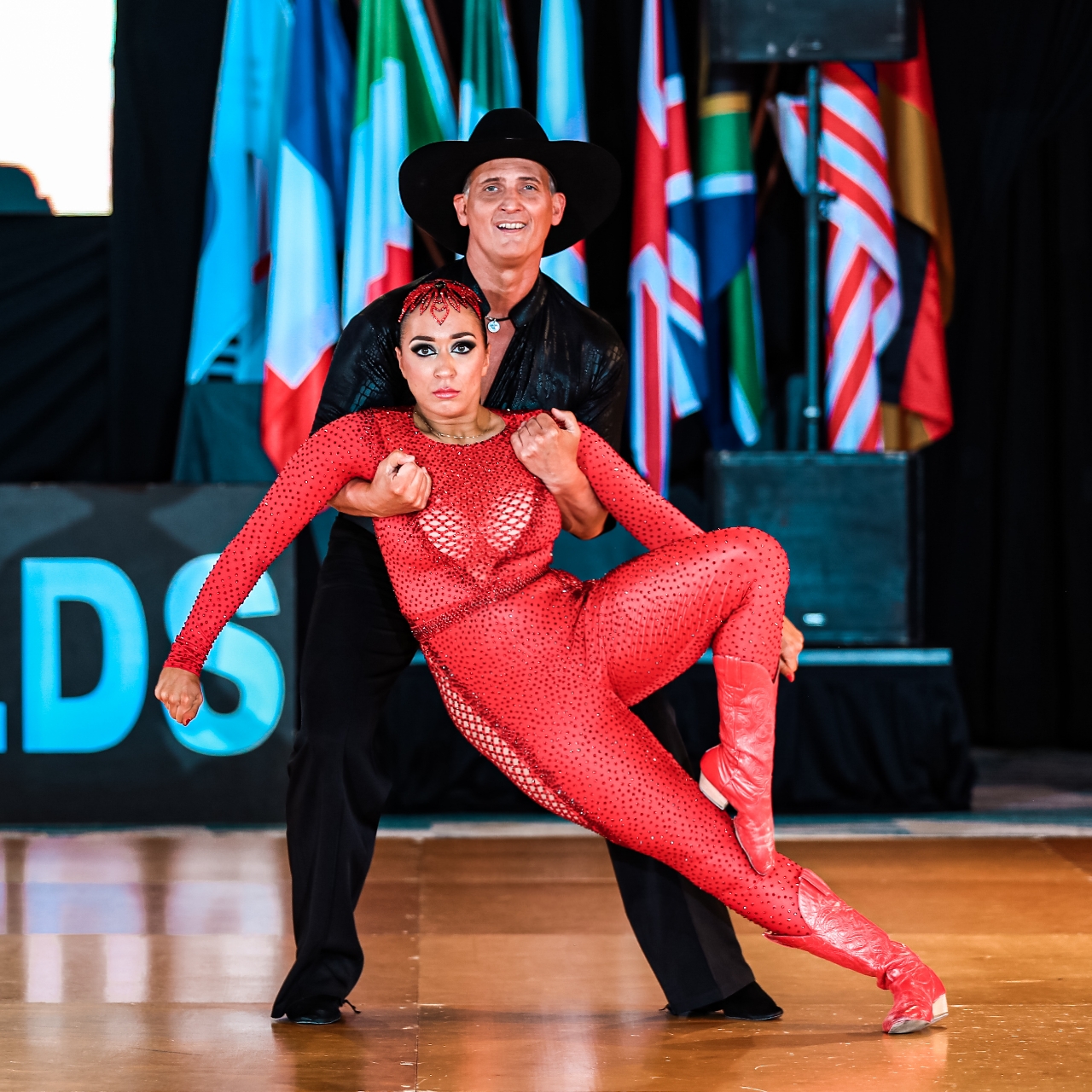 A couple dancing on a wooden floor with international flags in the background, the woman in a red outfit and the man in black.