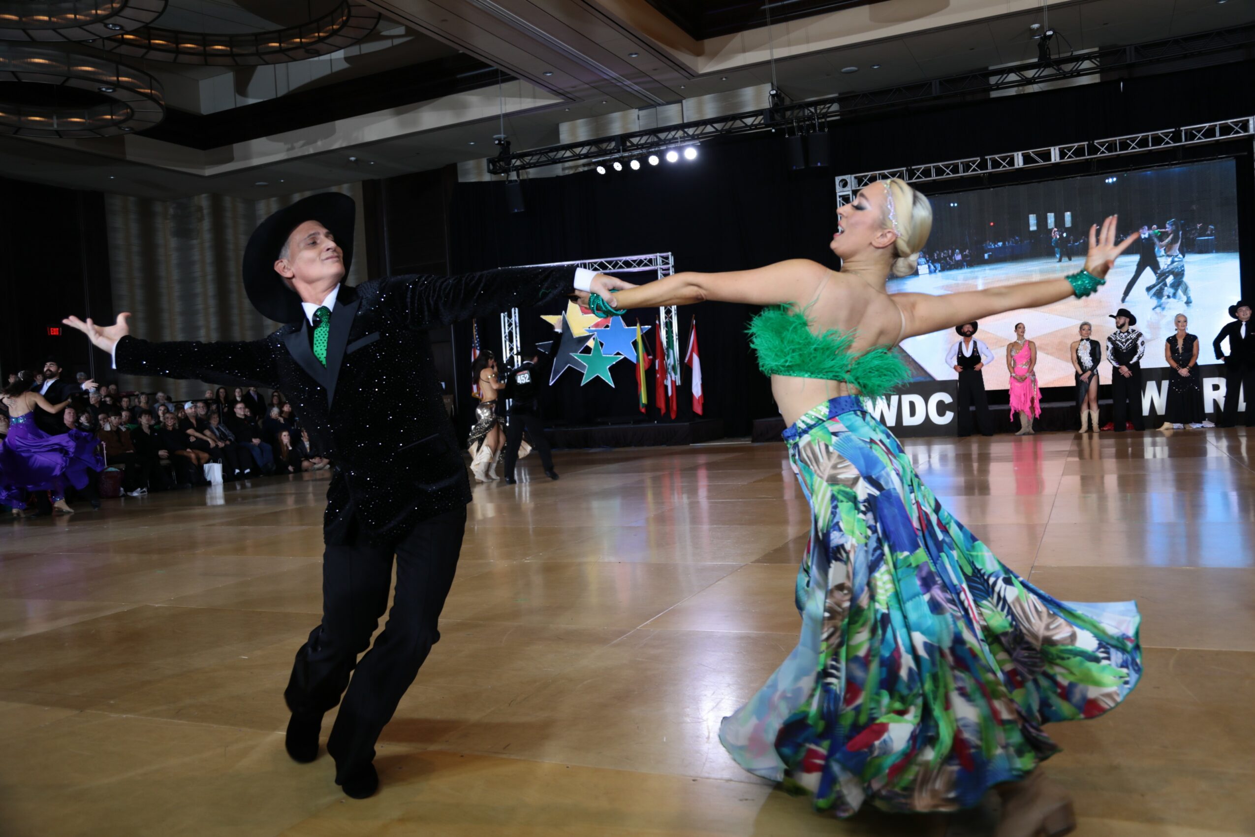 A couple dancing on a ballroom floor, with the woman in a colorful skirt and the man in a black suit, audience in background.