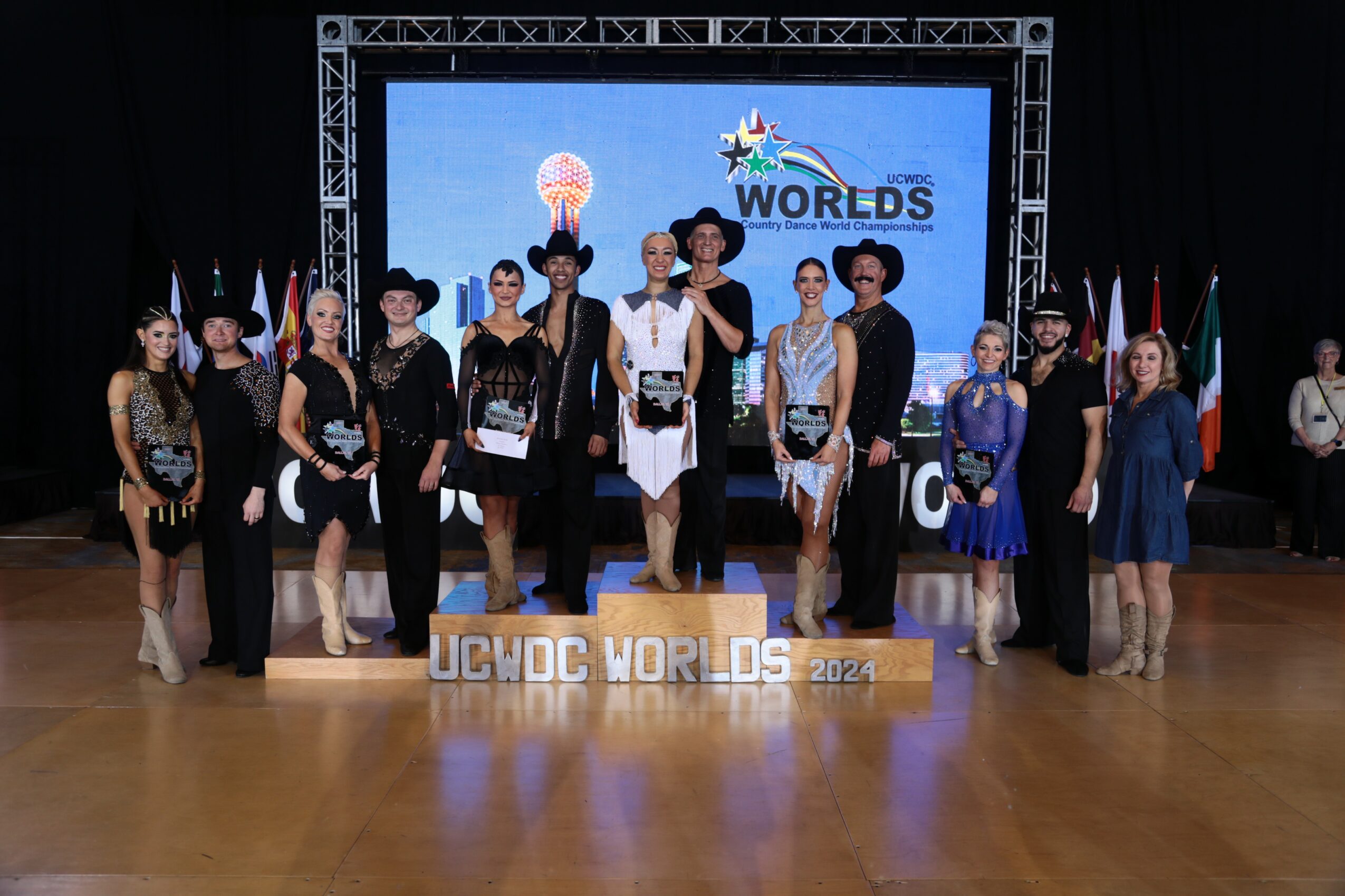 Group of people standing on stage with a banner reading 'JUNIOR WORLDS' in front, background shows a screen with a globe and text.