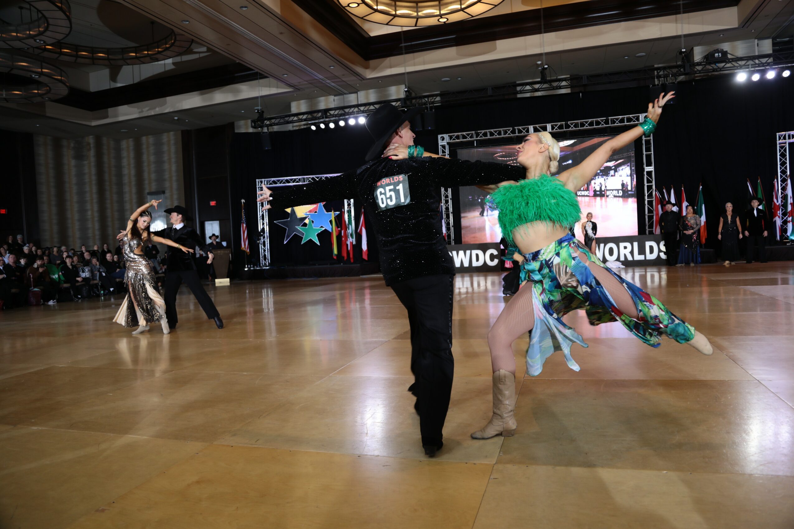 Two couples dance on a ballroom floor with a large screen and audience in the background, one couple in colorful costumes.