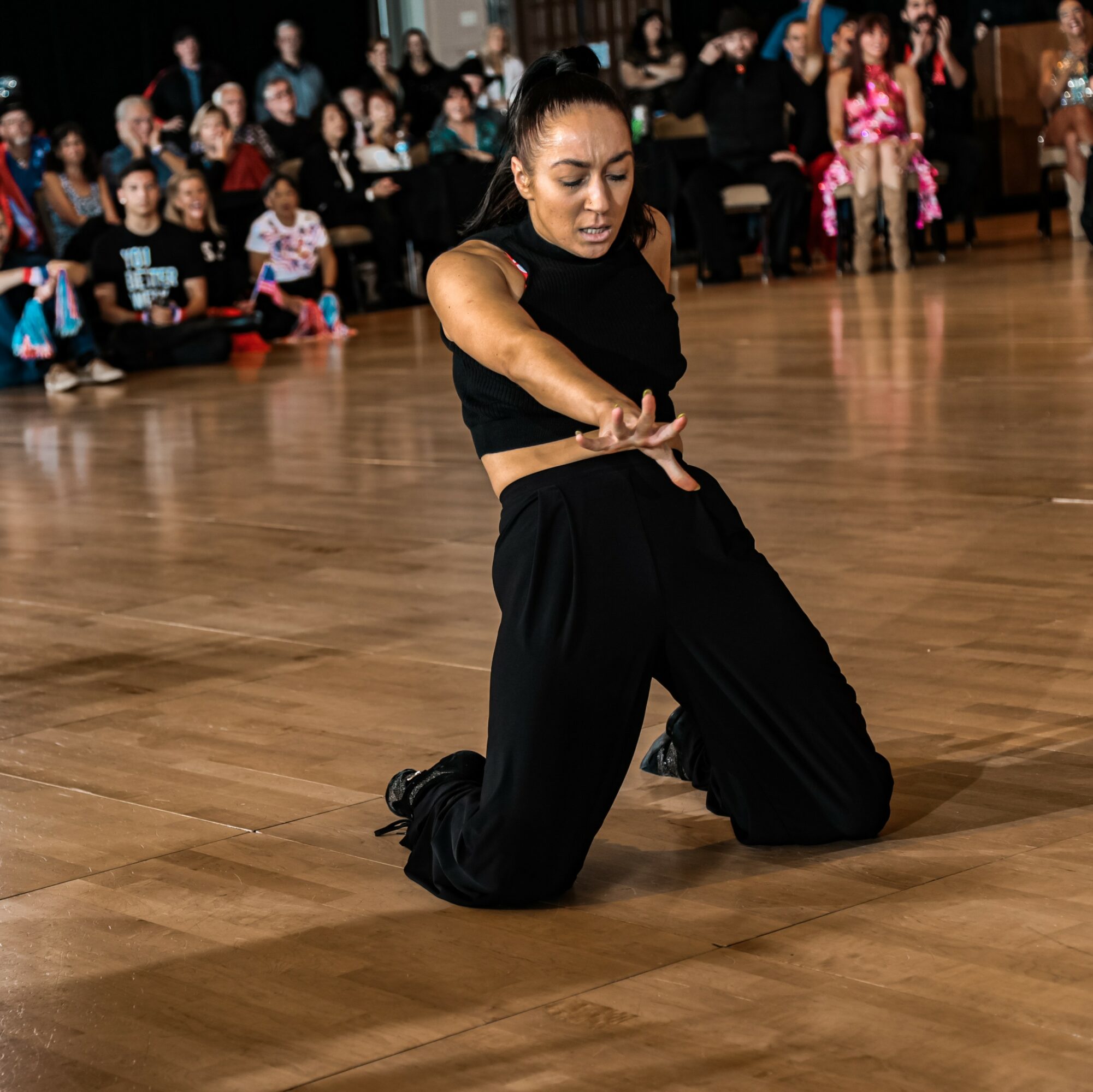 Woman kneeling on a dance floor with an audience watching in the background.
