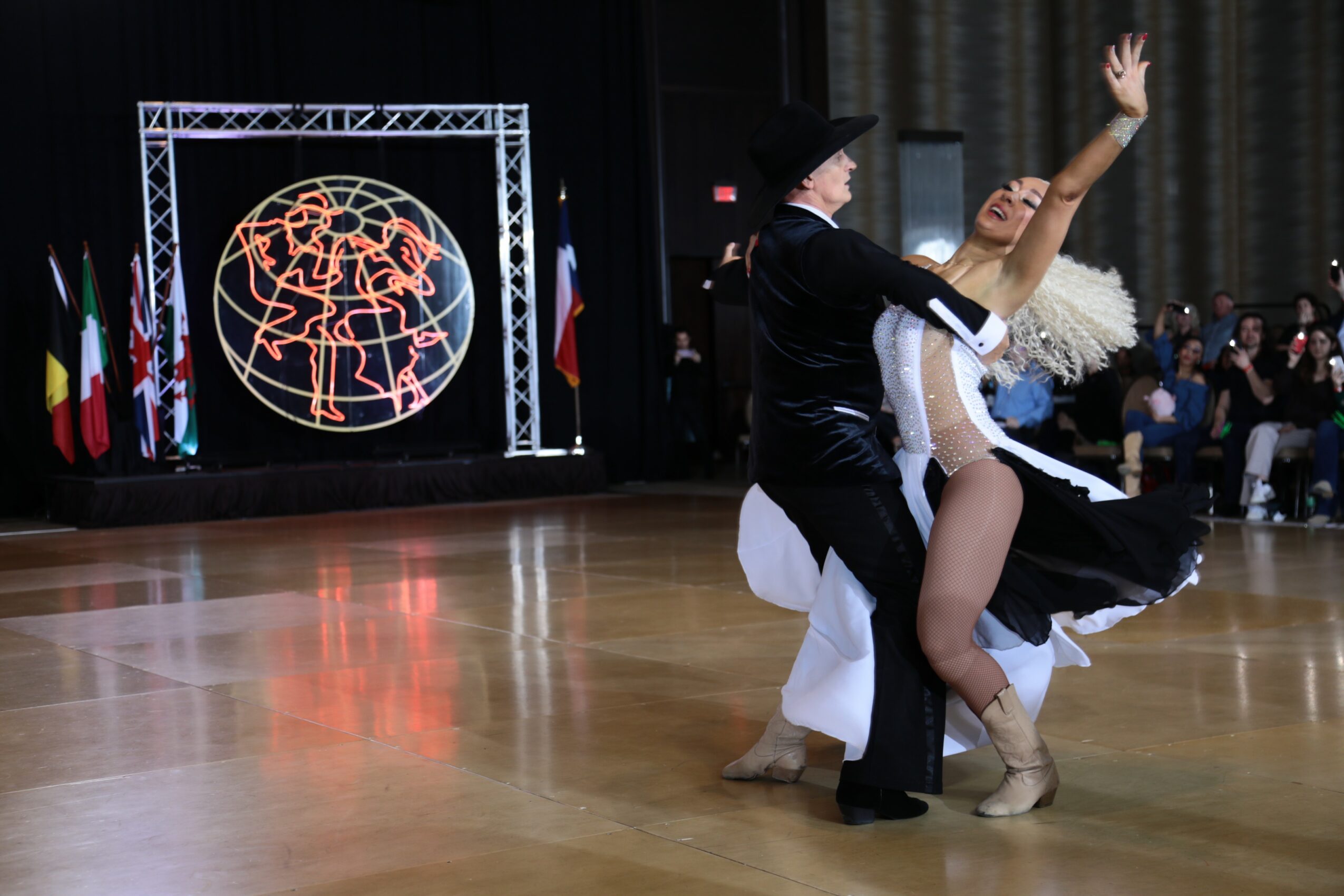 A couple dancing ballroom style on a polished floor with an audience and a digital globe display in the background.