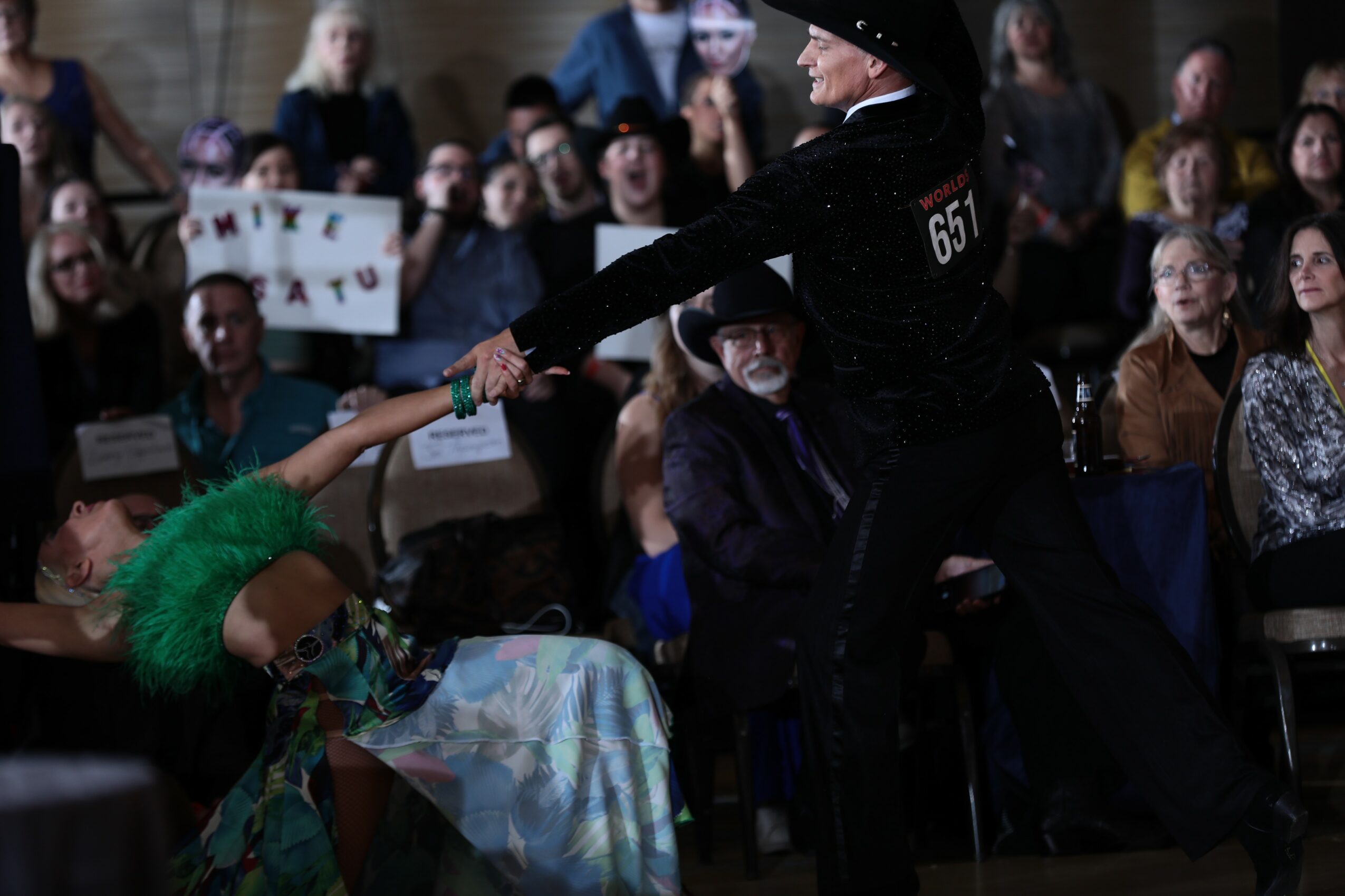 A person in a black outfit and hat hands a green feathered item to a seated person with a colorful dress, surrounded by an audience.