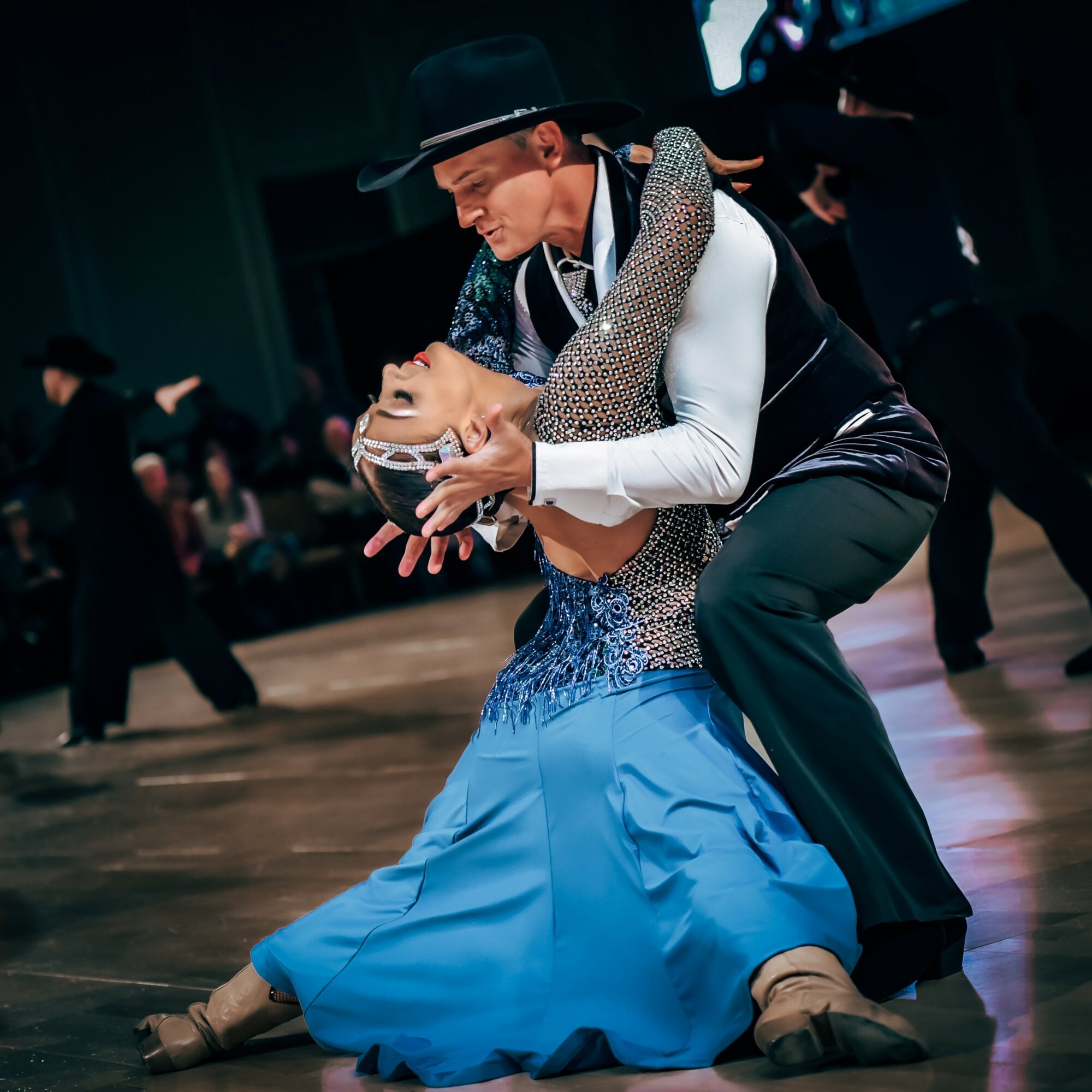 A couple dancing, the man wearing a black hat and vest, the woman in a blue dress, performing a ballroom dance.