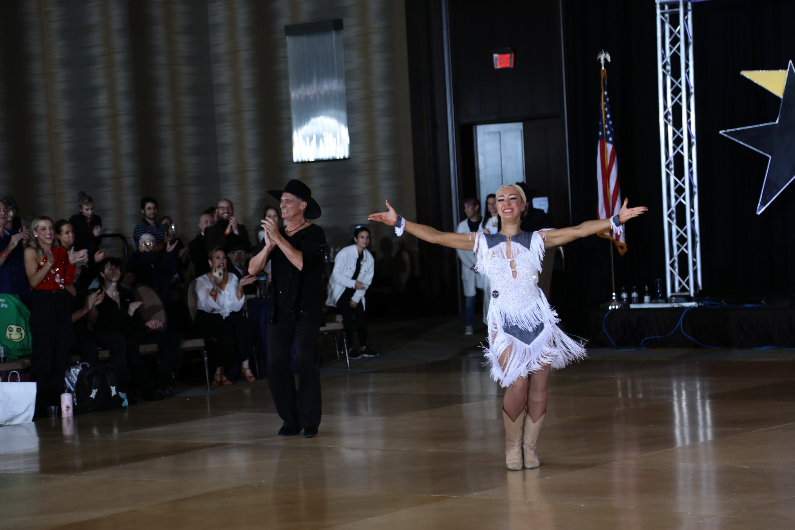 A woman in a white dress with arms outstretched performs on a dance floor, with an audience and American flag in background.