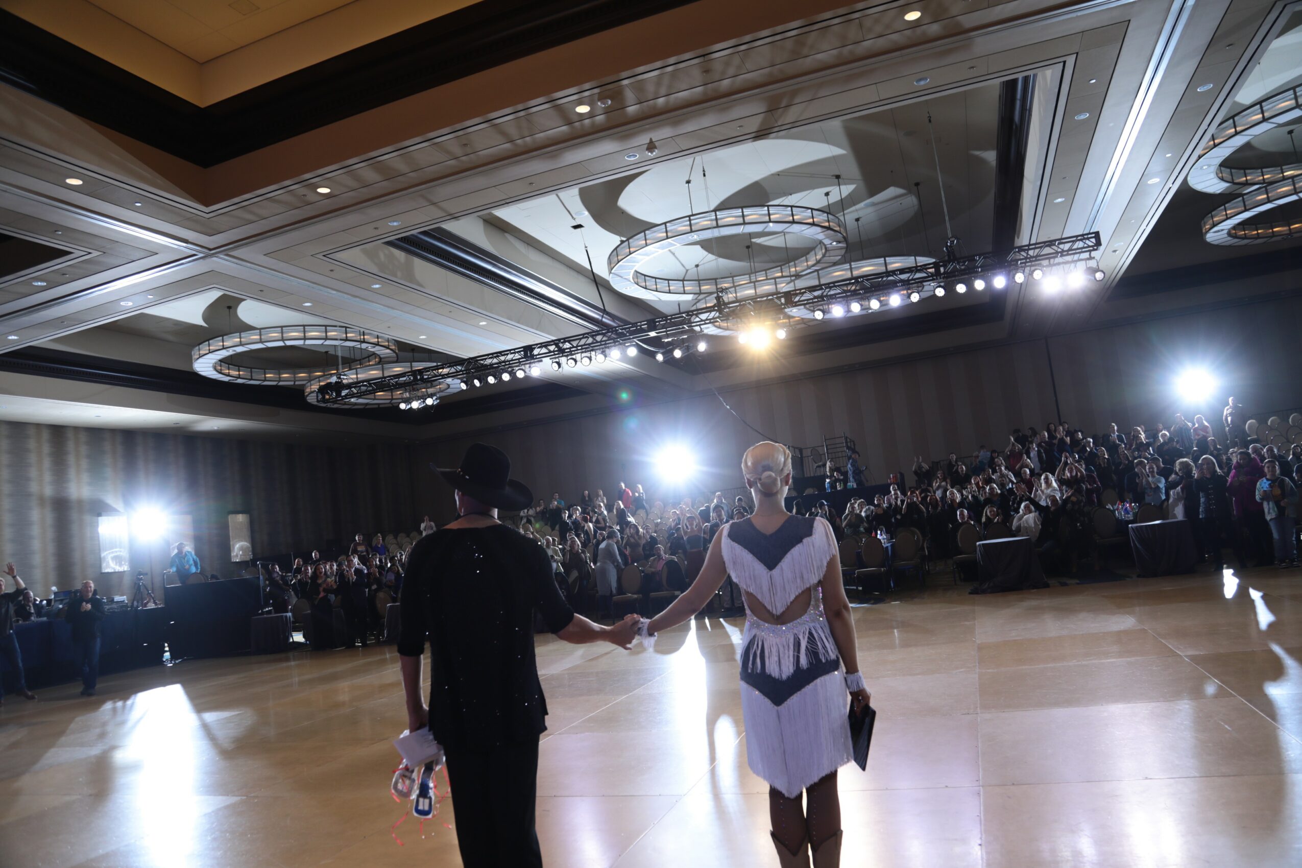 Two people dance in a large ballroom with an audience and bright overhead lights, holding hands and facing each other.