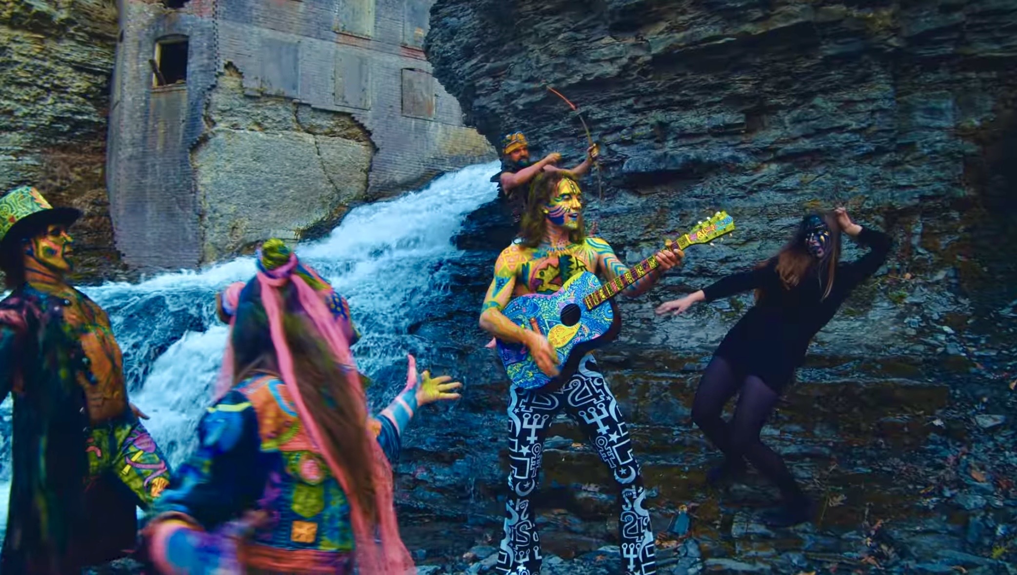 Group of people in colorful clothing near a waterfall, one playing guitar, others dancing or posing outdoors.