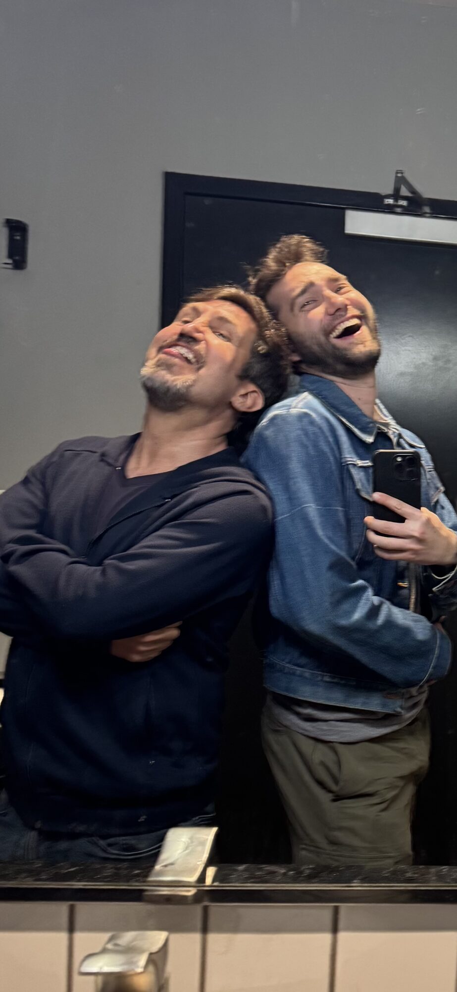 Two men smiling and laughing, leaning against a mirror in a room with a dark door behind them.