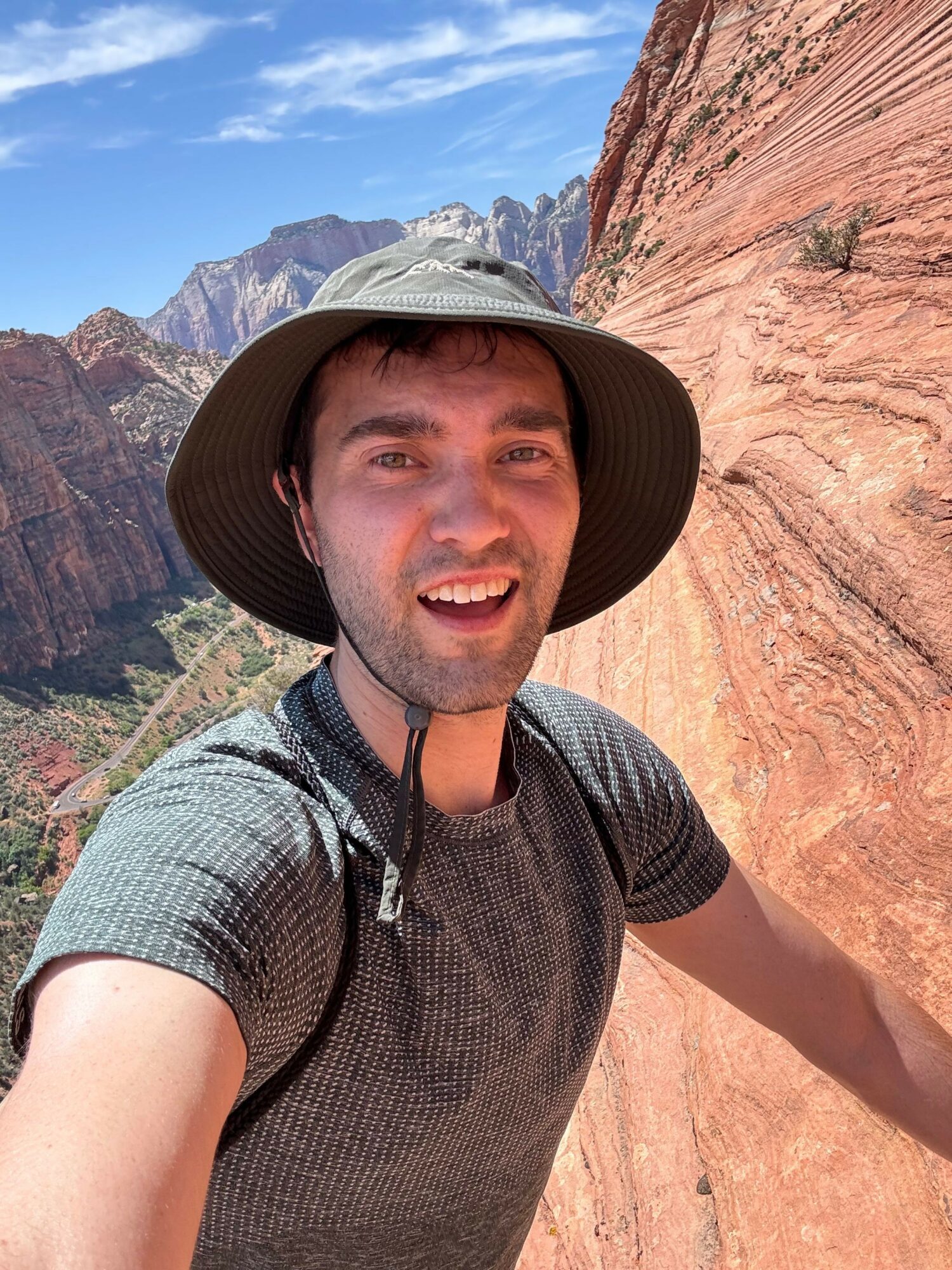 Person smiling, wearing a wide-brimmed hat, taking a selfie in a desert landscape with red rock formations and blue sky.