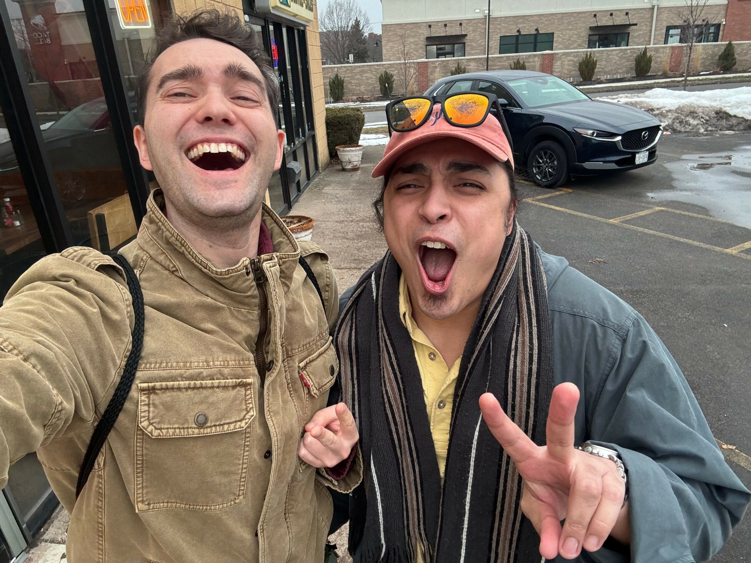 Two people taking a selfie outdoors, one making a peace sign, both smiling with open mouths, in front of a building and parked cars.