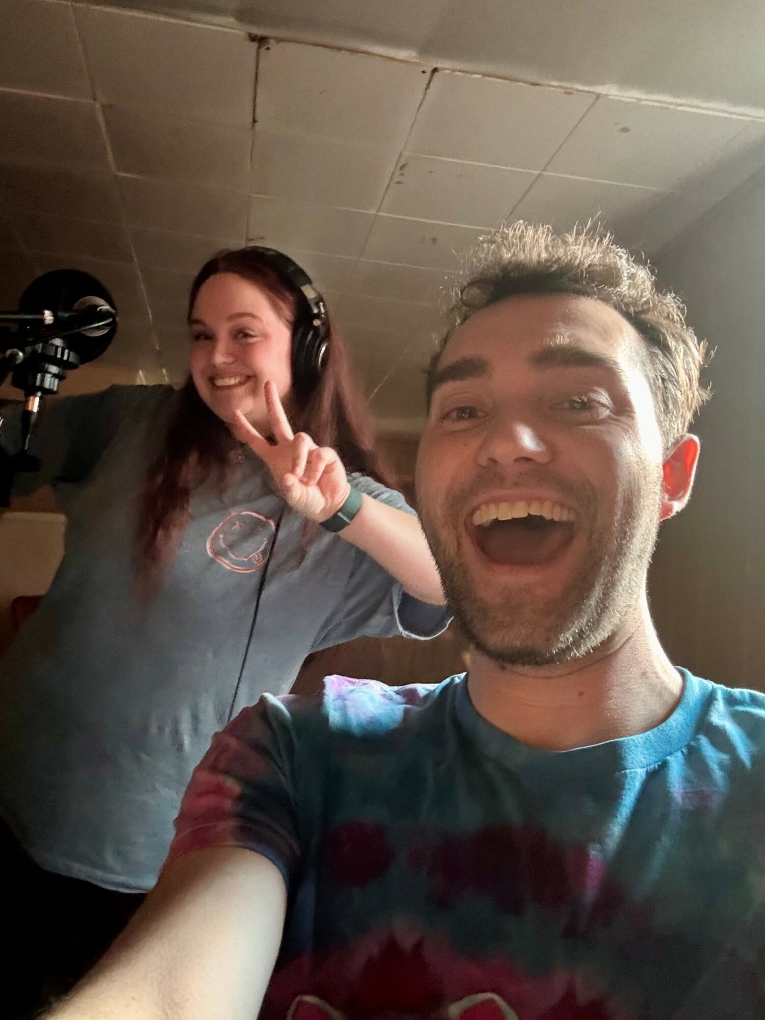 Two people smiling, one making a peace sign, in a recording studio with a microphone, casual clothing, and headphones.