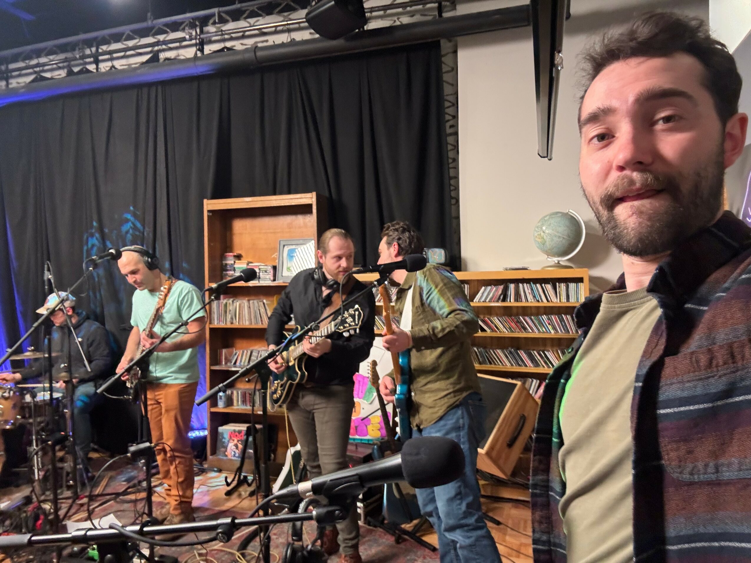 Group of musicians performing on stage with bookshelves in the background, one person taking a selfie, and musical instruments visible.