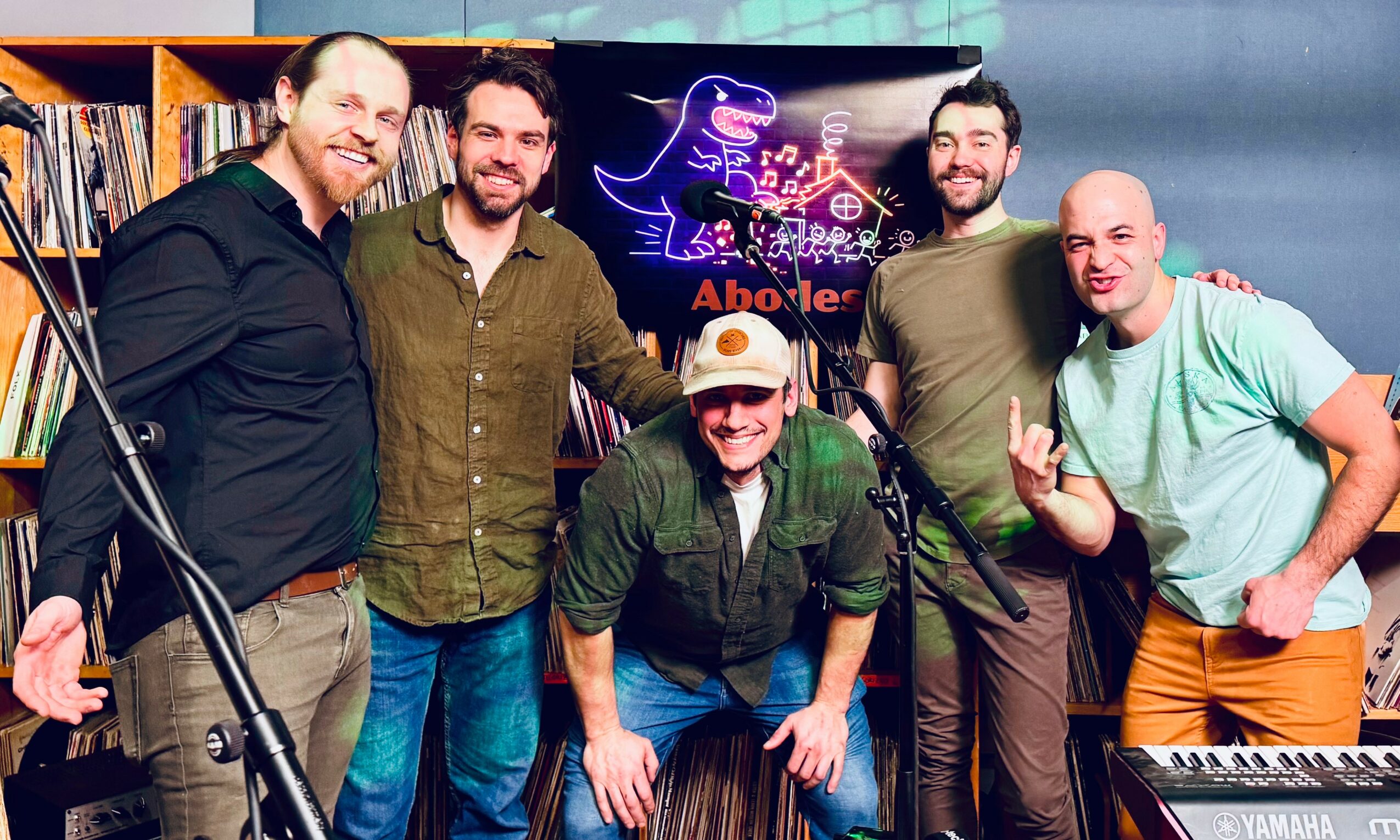 Group of five men smiling, standing in a room with musical equipment and a neon sign in the background.