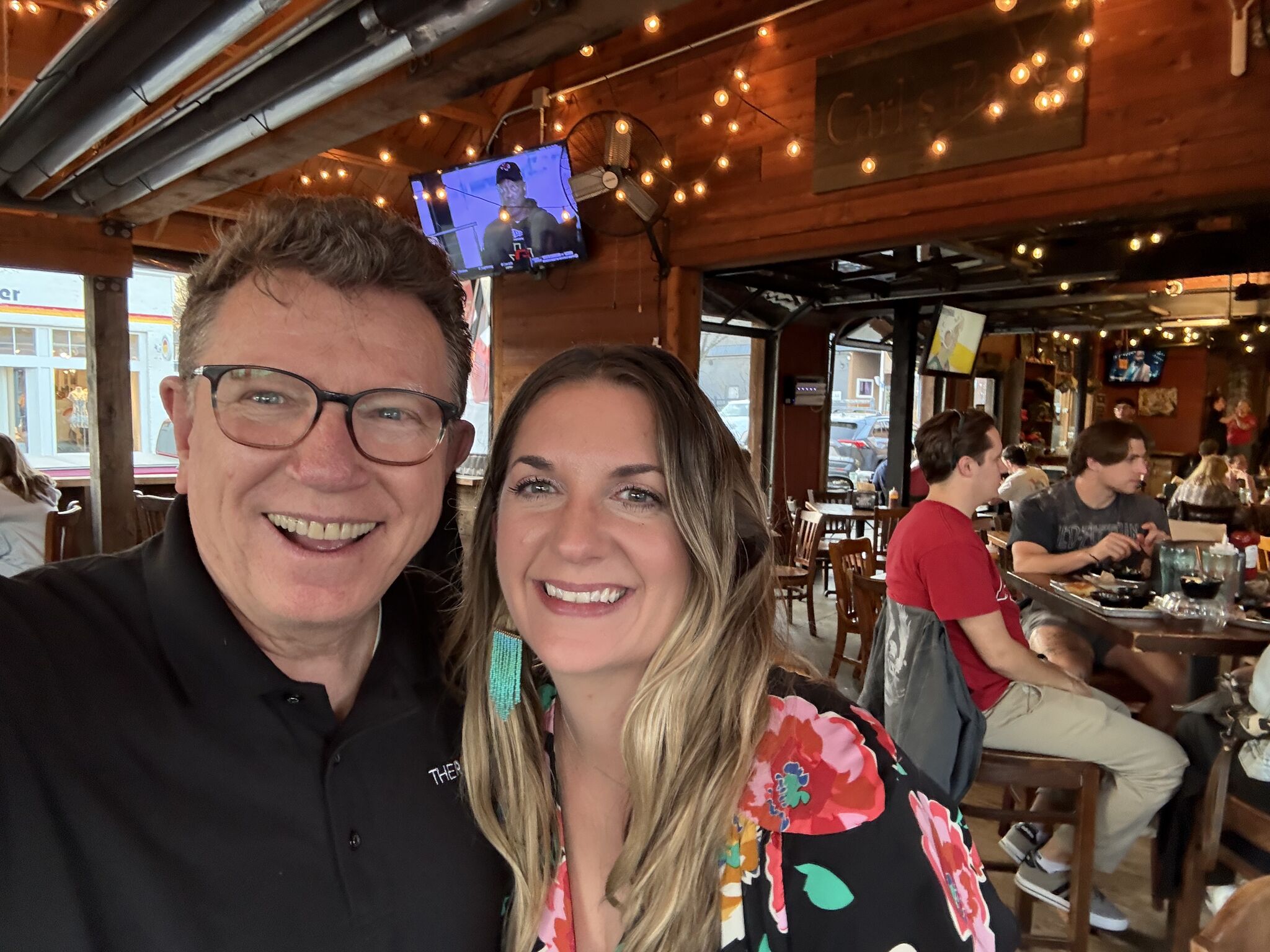 Two people smiling in a restaurant with string lights and a TV in the background.