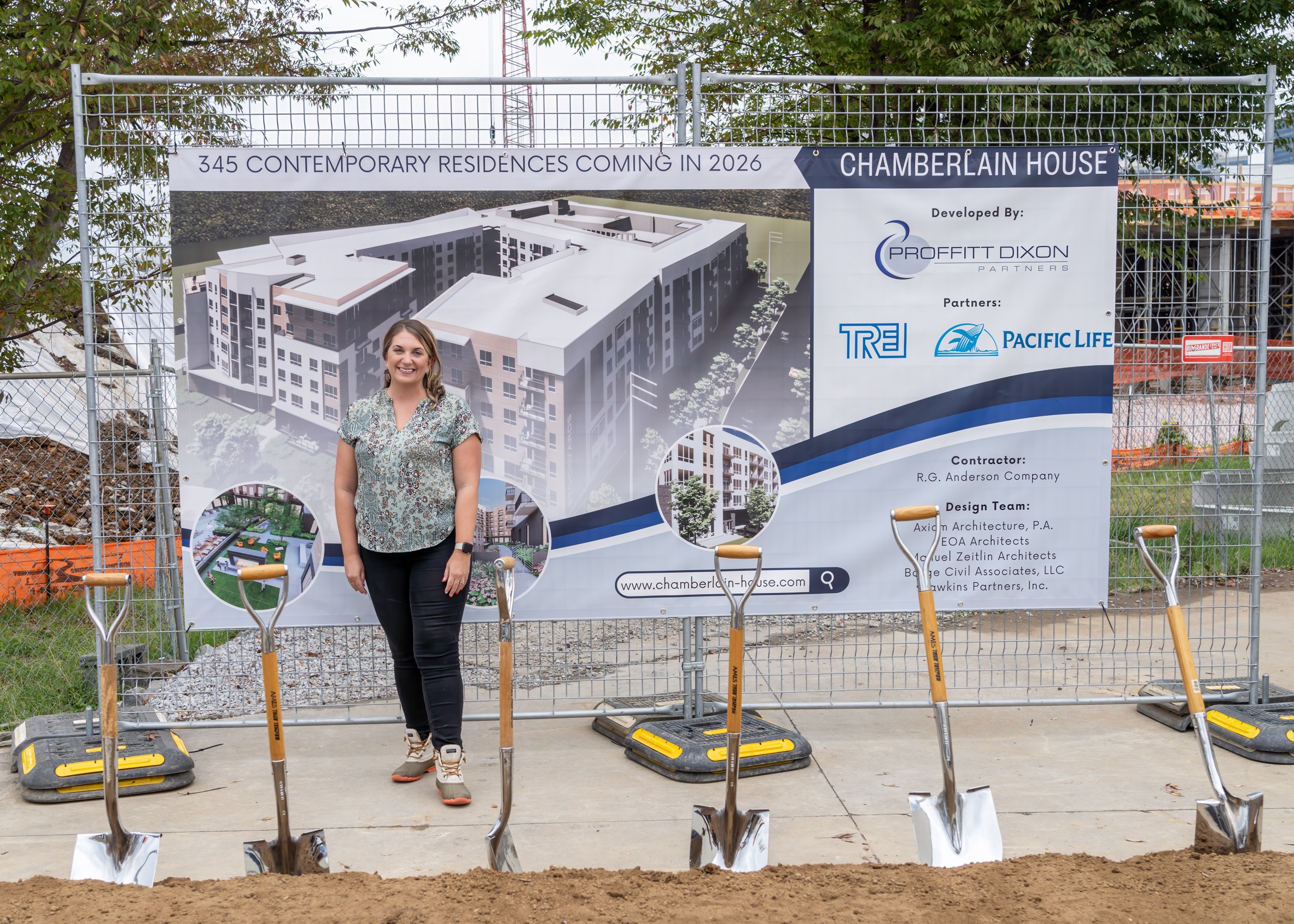 Woman standing in front of a construction site with a large banner showing an architectural rendering of a building, trees, and construction tools.