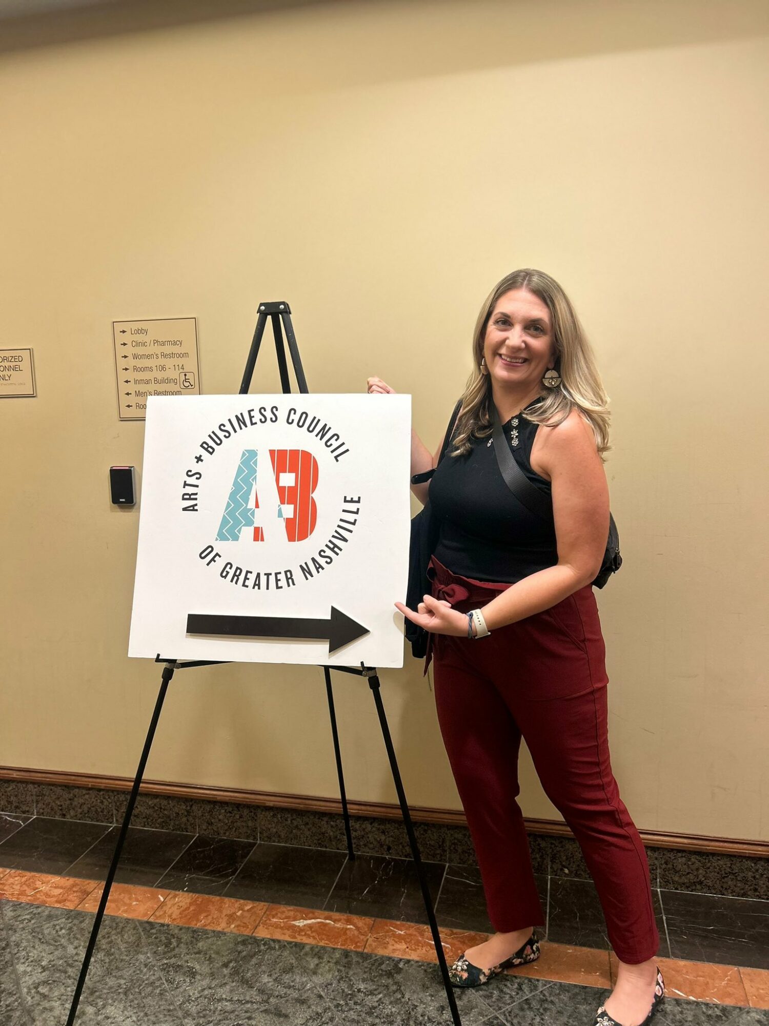 Woman standing next to a sign with the AB logo and an arrow, smiling, in an indoor setting.